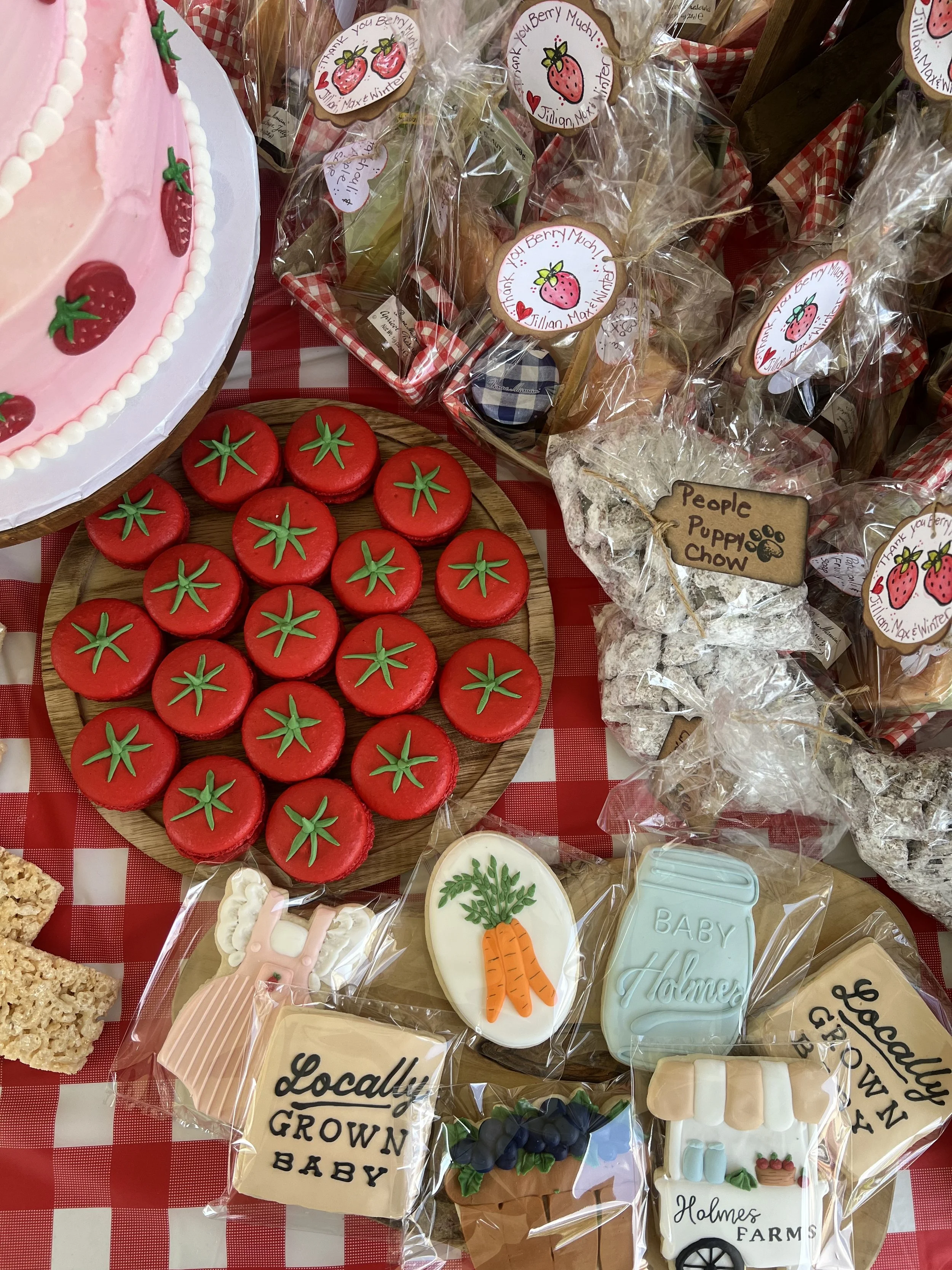 Decorative cookies shaped like tomatoes, baby carrots, a baby bottle, and related farm and baby themes, along with a pink cake with strawberries and a box of baked goods with strawberry and farm-themed labels on a red and white checkered tablecloth.