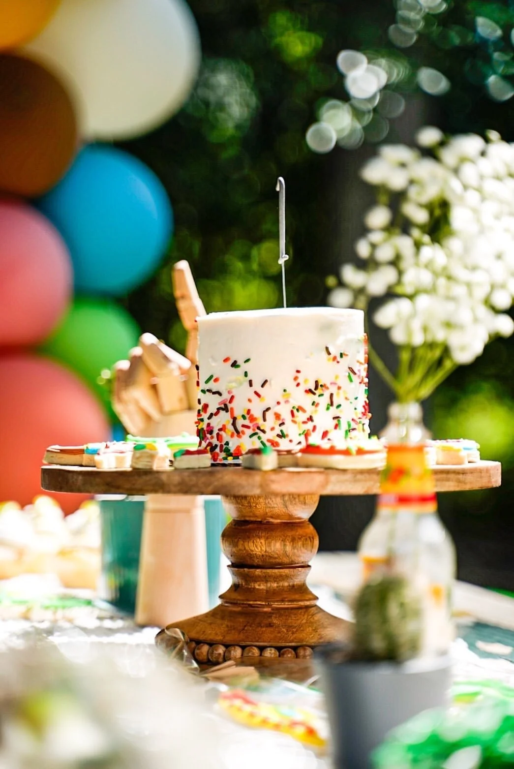 A birthday cake with white frosting and colorful sprinkles on a wooden cake stand, surrounded by cookies, with balloons and white flowers in the background.