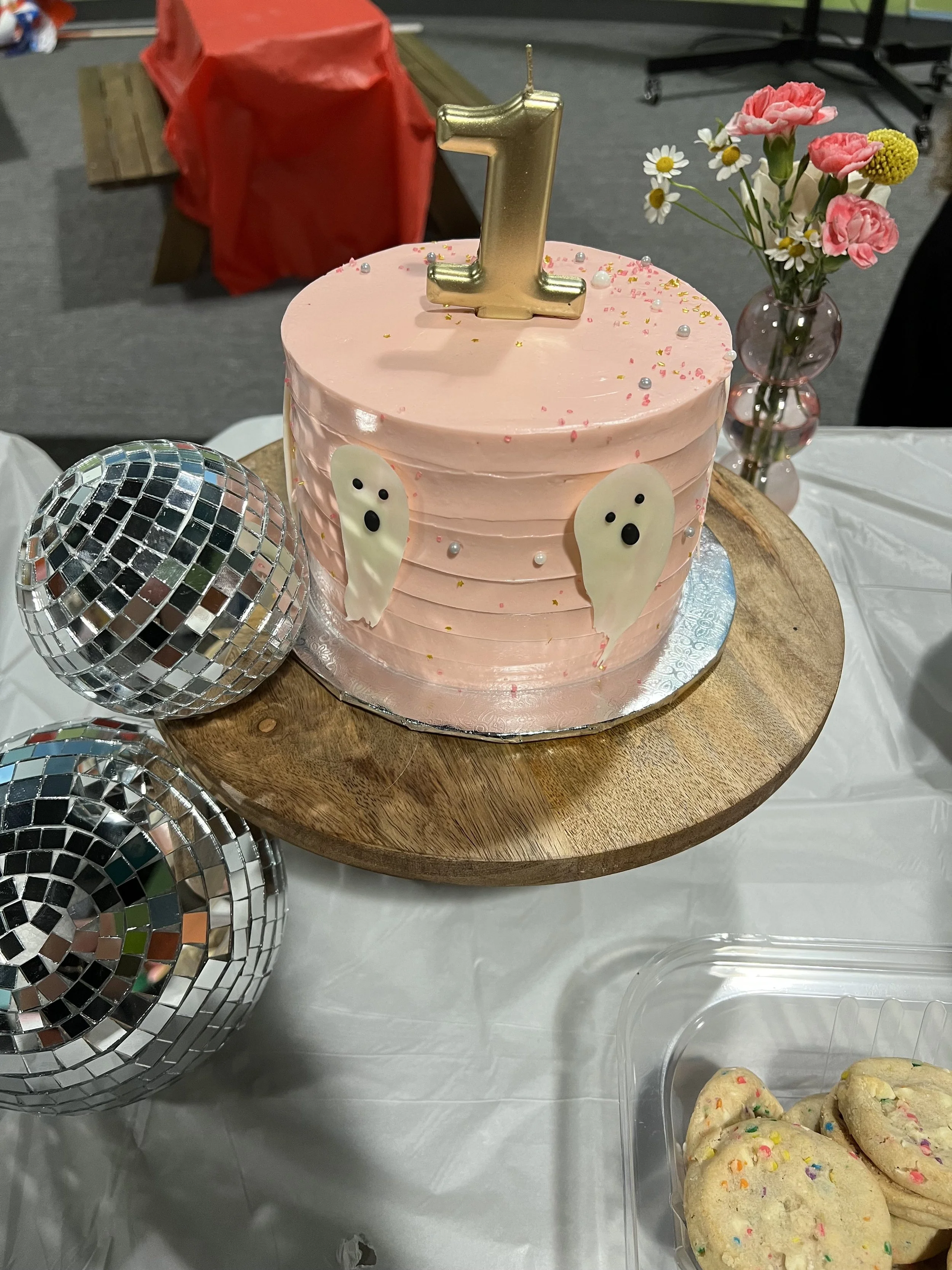 Pink birthday cake with a gold number one candle on top, decorated with white ghost shapes and small colorful sprinkles, placed on a wooden cake stand. Decorative daisies and pink flowers in a vase are in the background, with disco balls and cookies