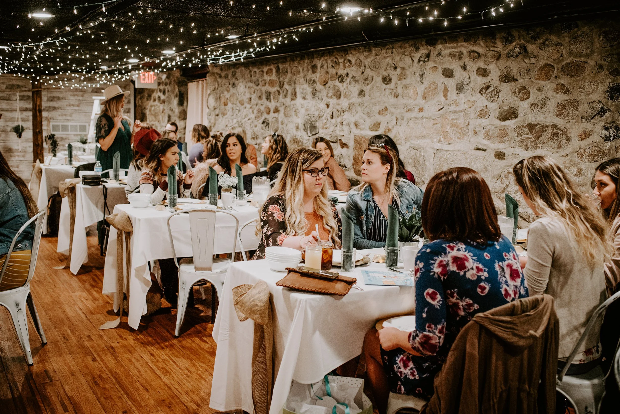 Women sitting at decorated tables with white tablecloths, green napkins, and candles, in a rustic venue with string lights and a stone wall, at a social event or gathering.