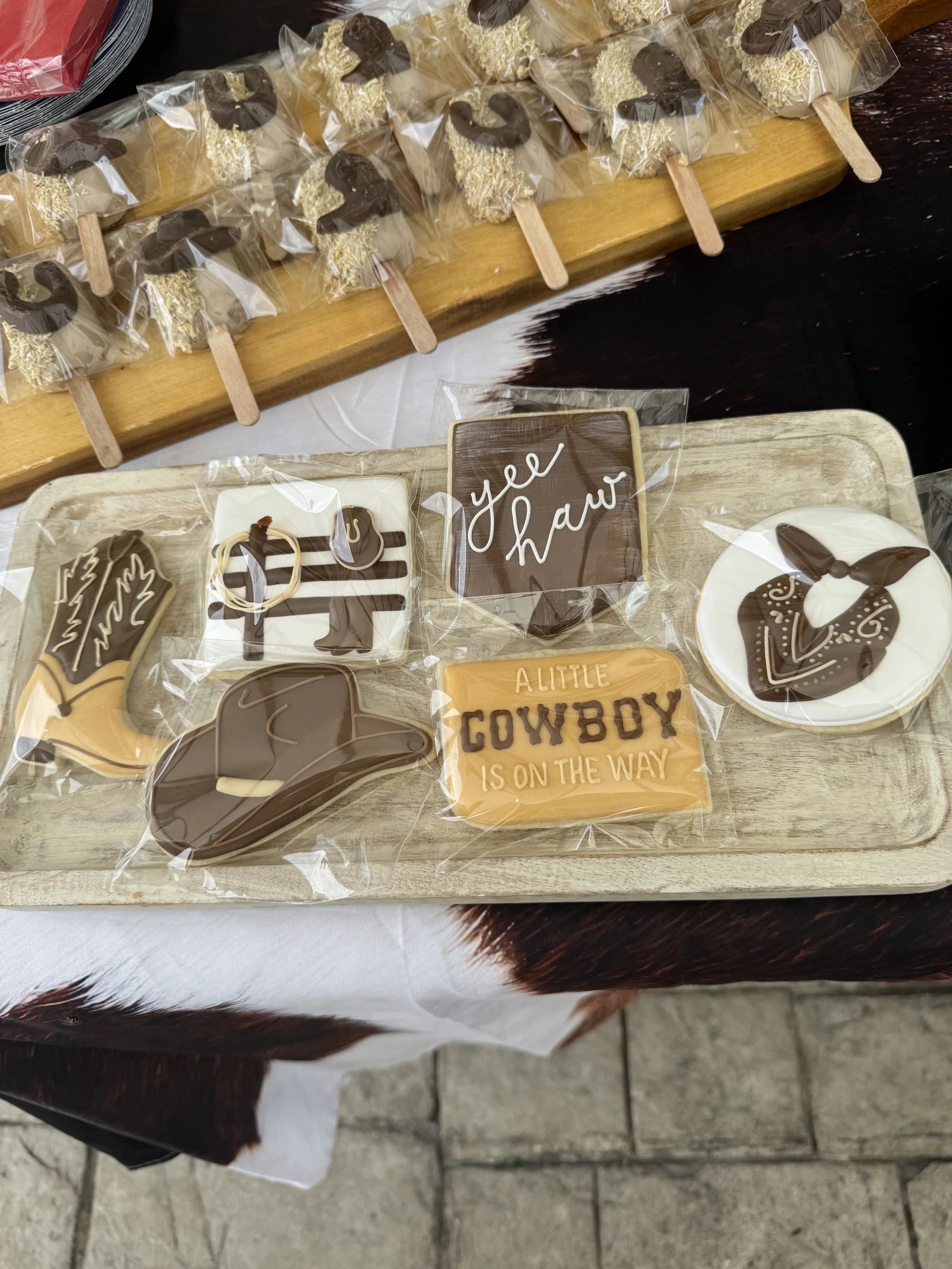 Assorted cowboy-themed cookies, including boots, hats, and a sheriff badge, along with a cookie that says 'Yeehaw' and another that reads 'A little cowboy is on the way,' all covered in plastic wrap.