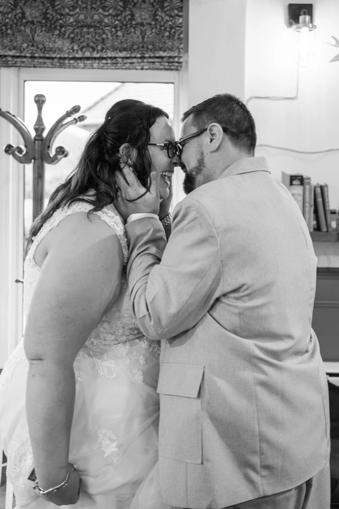 A couple is sharing an intimate moment, touching foreheads and smiling with noses almost touching, in a room with a window and bookshelf in the background.