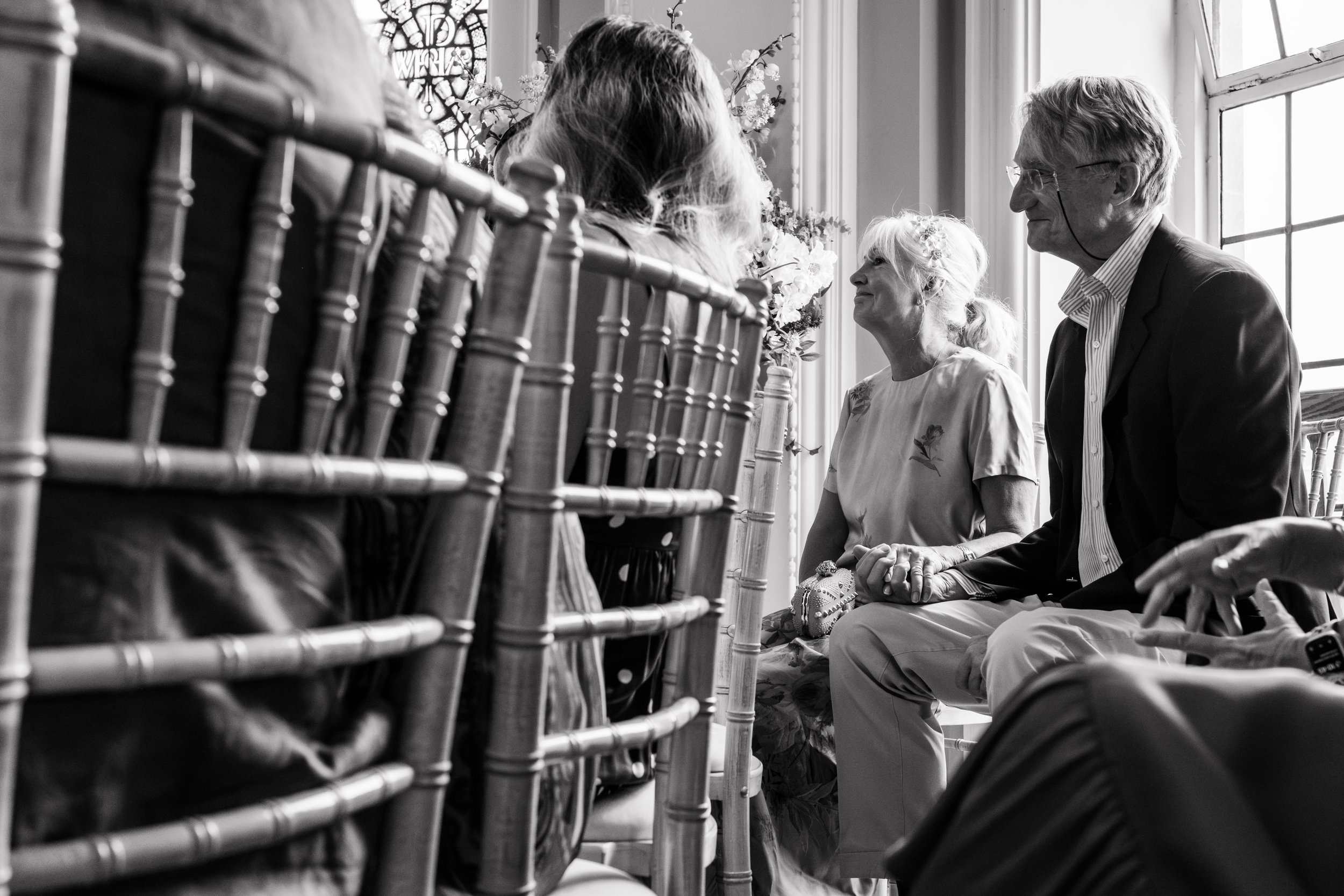 A black-and-white photo of a wedding ceremony with three people sitting in chairs near a window. A woman in a light-colored blouse and a man in a suit are holding hands and smiling at a woman across from them. The scene is decorated with flowers and 