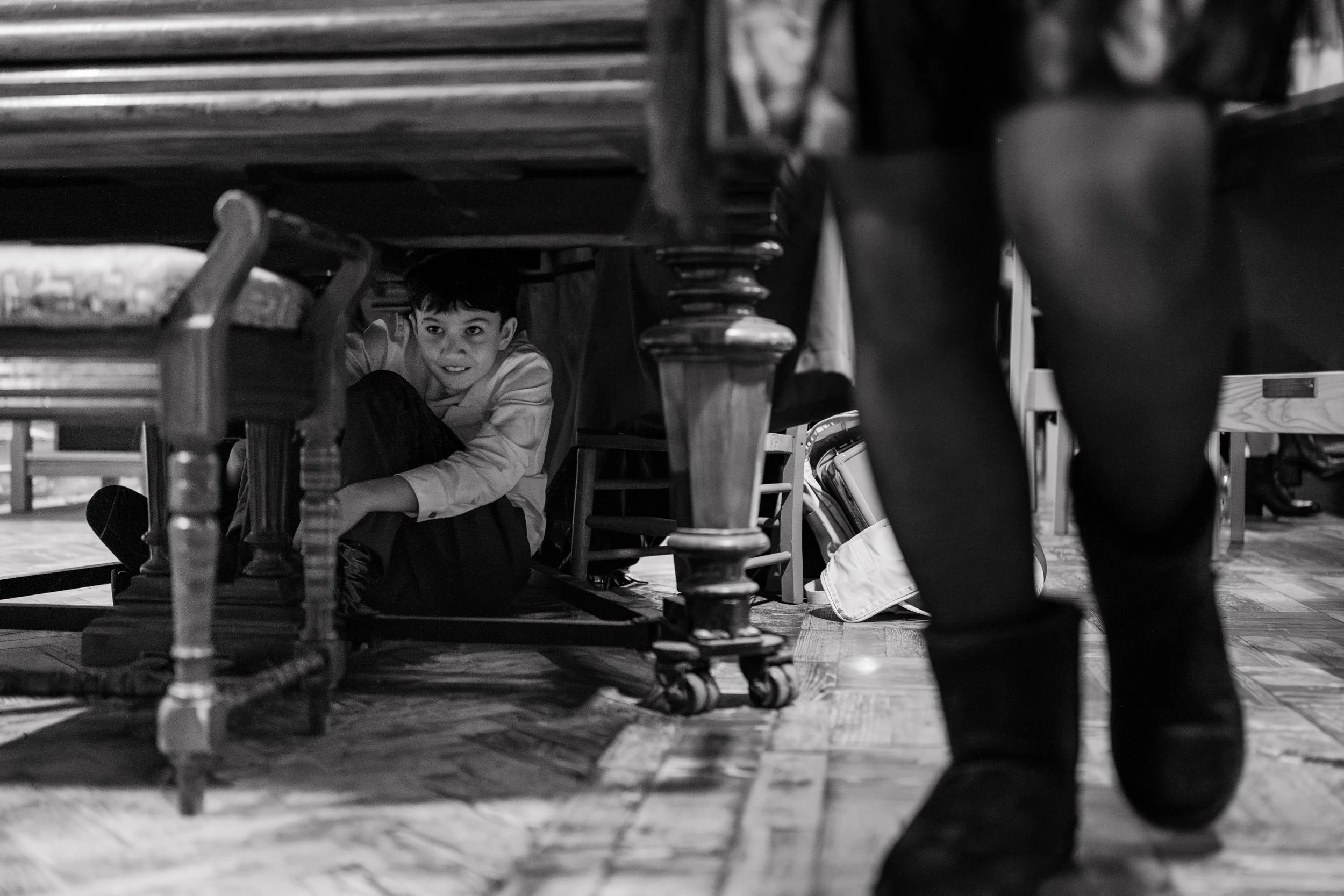 A black and white photo of a boy hiding under a table, looking out with a smile, while an adult stands nearby on a wooden floor.