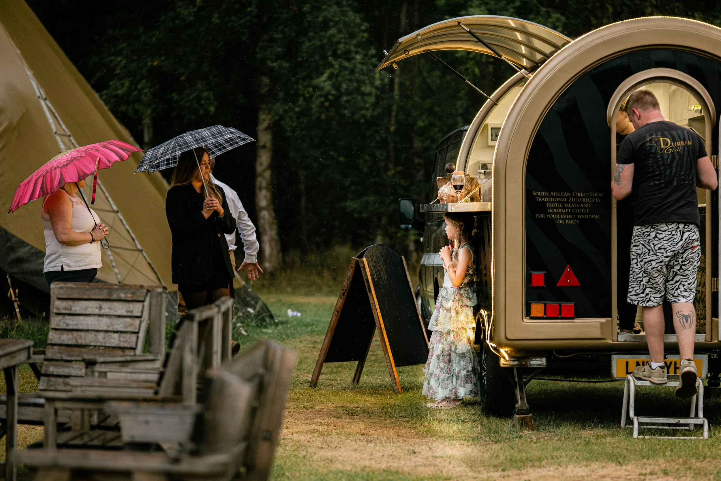 People with umbrellas standing outside near food truck and a young girl in a floral dress eating ice cream on a rainy day in a park.