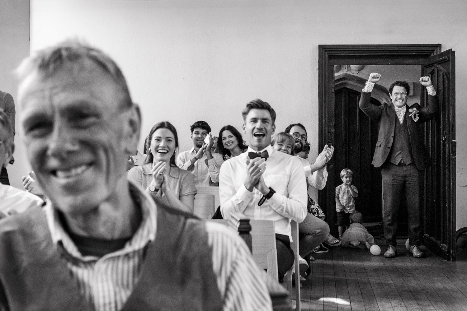 A black-and-white photo of a joyful group of people at a wedding or celebration, clapping and smiling, with a man in a suit and bowtie sitting in the front. In the background, a person with glasses and a patterned shirt is raising their fists in exci