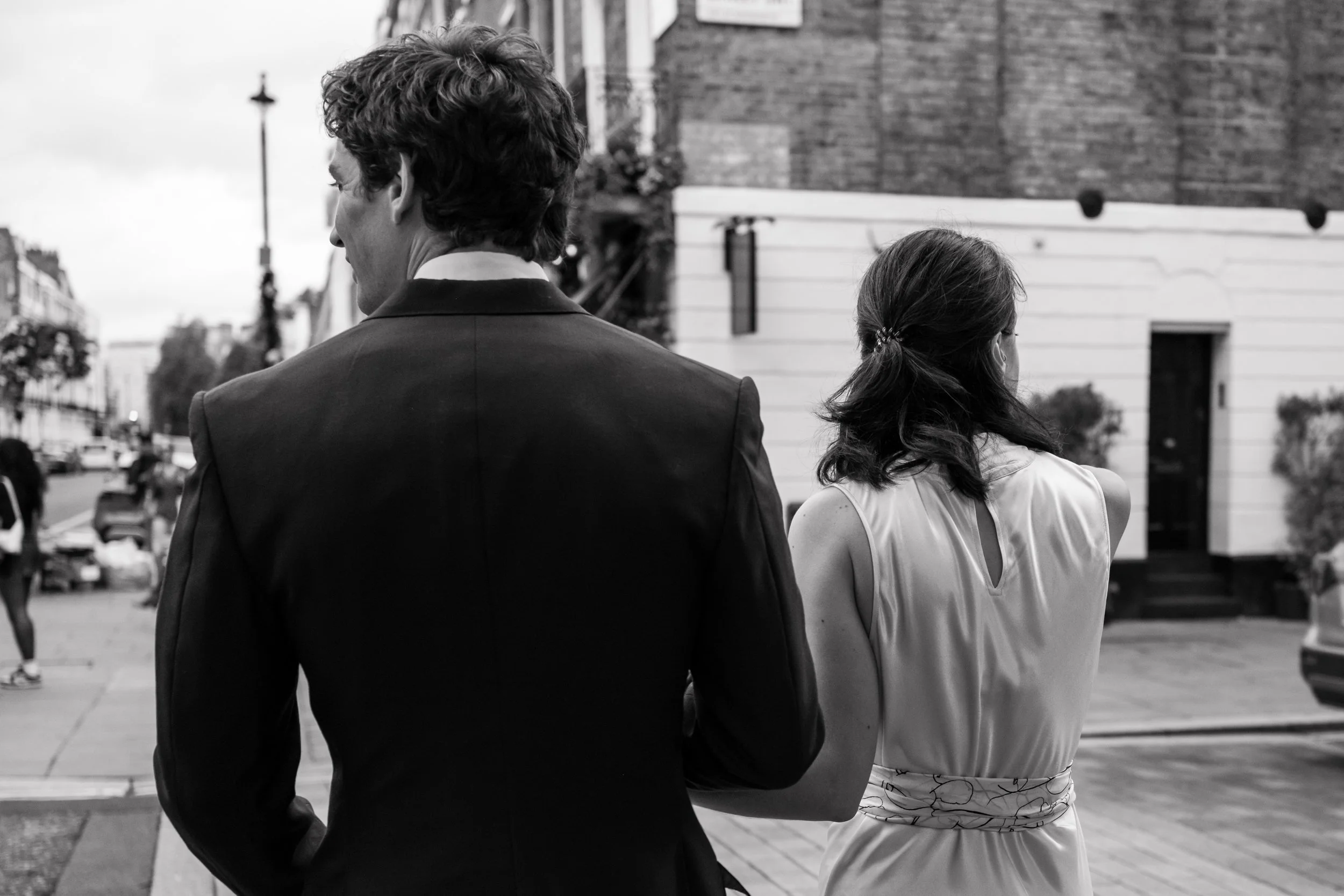 A black and white photo of a man and woman walking outside on a city street, seen from behind. The man is wearing a suit, and the woman has dark hair tied back and is wearing a sleeveless dress.