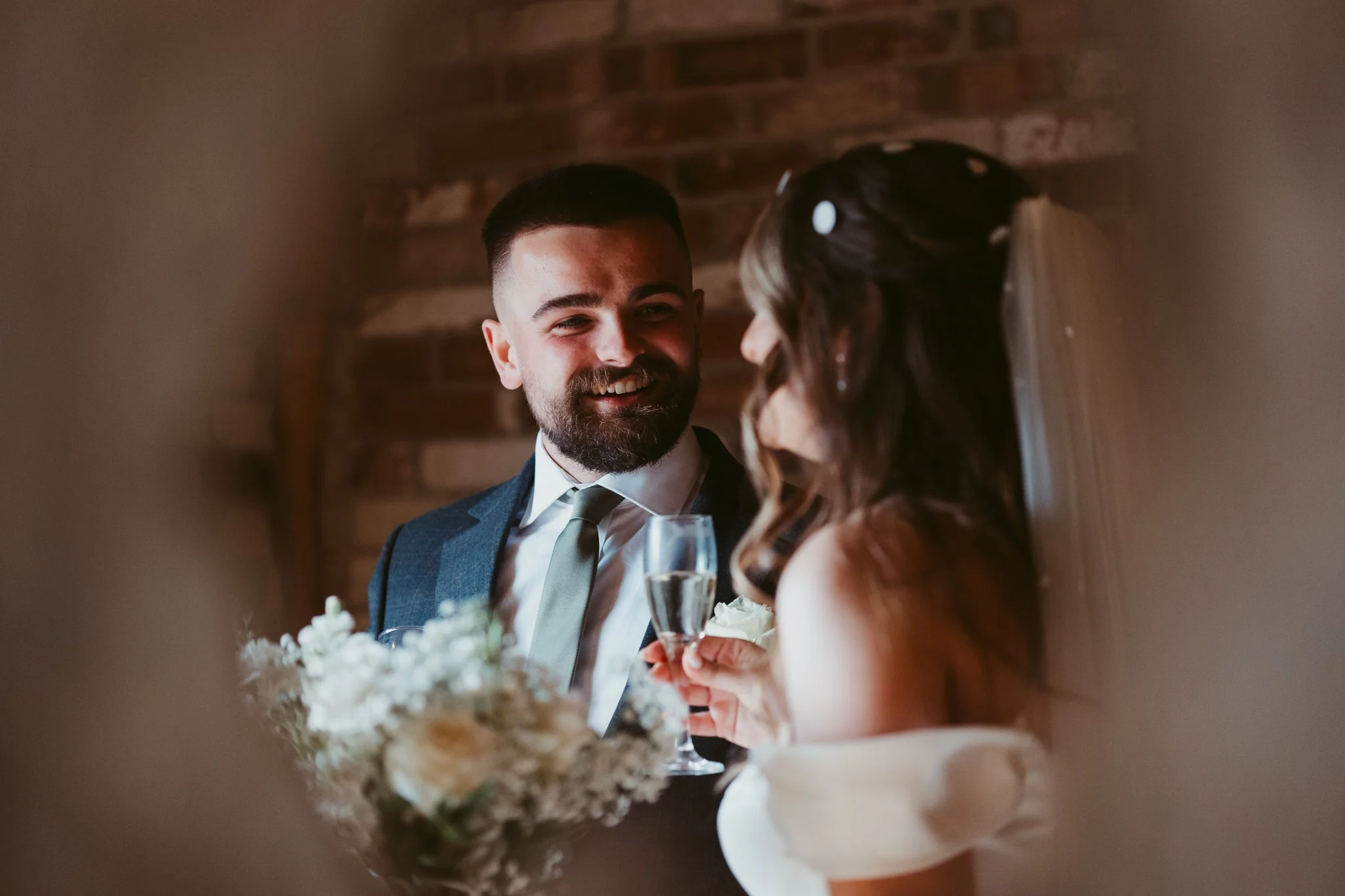 A man and woman sharing a toast at a wedding reception, with the man smiling and the woman holding a glass of champagne, floral bouquet in the foreground, brick wall background.