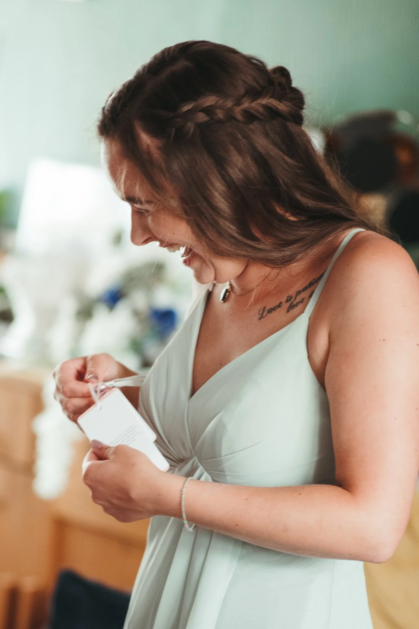 A woman with brown hair styled in a braid, wearing a light-colored dress, looking at an item in her hands with a smile.
