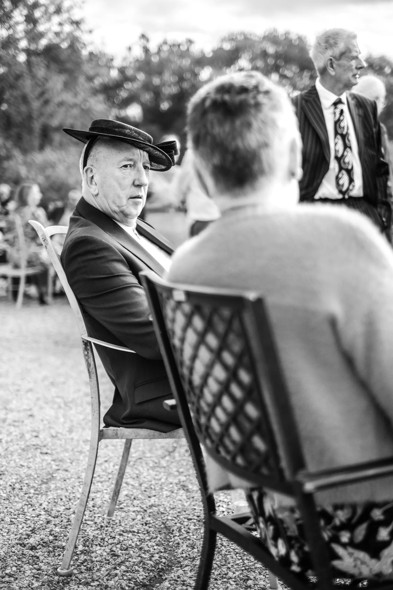 A black and white photo of a man wearing a suit and a decorative hat, seated outdoors at an event. He is looking at another person with short hair, sitting in front of him. In the background, a man in a striped jacket and tie stands, with trees and o