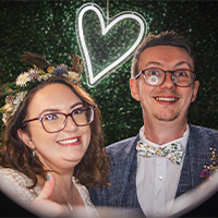 A smiling woman and man in formal attire, both wearing glasses, standing in front of a green leafy backdrop with a glowing heart outline above them.