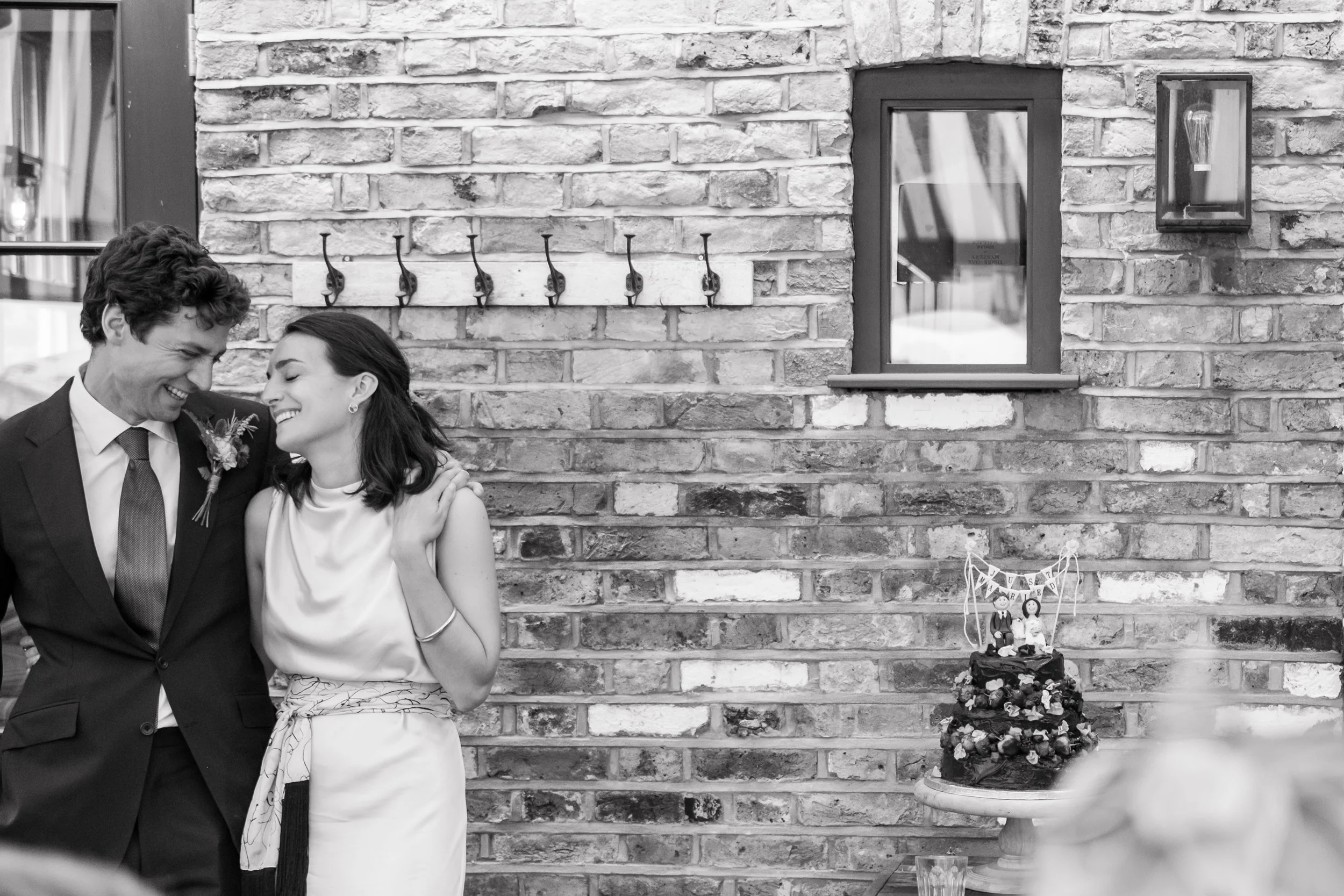 A bride and groom smiling and embracing at their wedding reception near a brick wall with hooks and a window, with a wedding cake on a stand to the right.