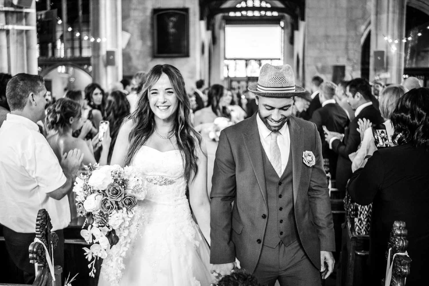 Black and white photo of a bride and groom walking down the aisle at a wedding ceremony inside a church, surrounded by guests.