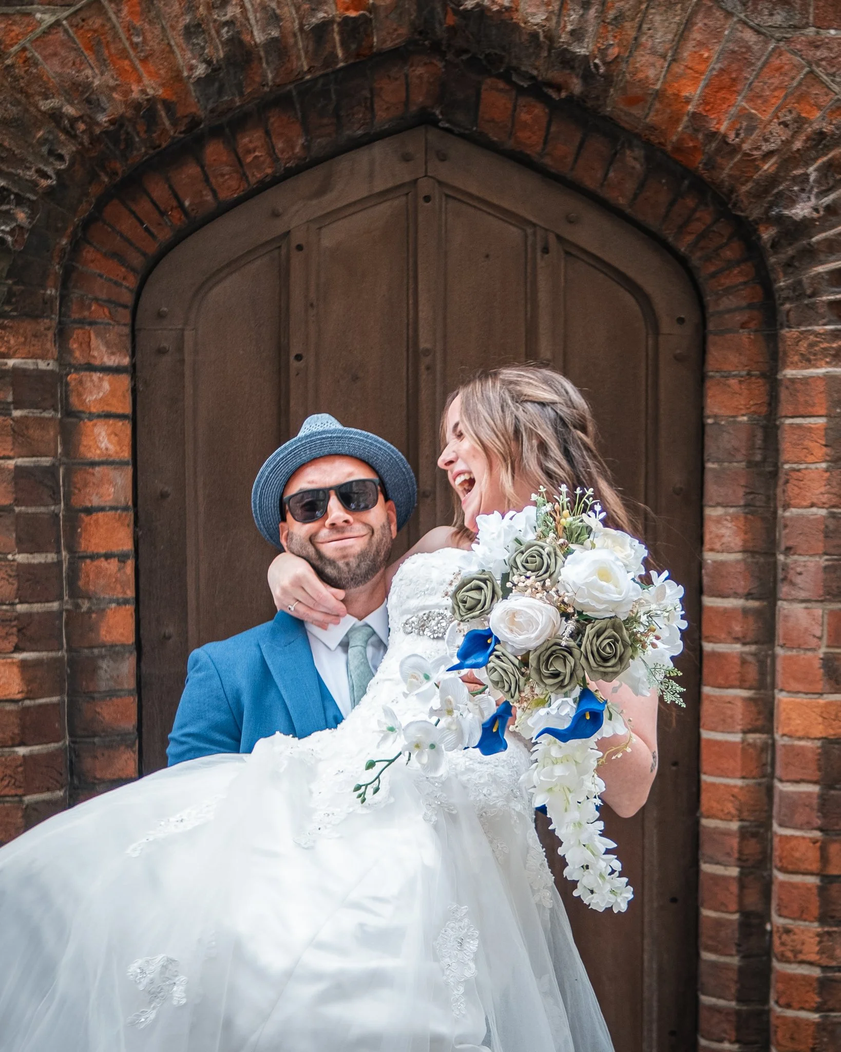 A bride and groom celebrating their wedding in front of a brick archway, with the groom in sunglasses and a blue suit, and the bride in a white gown holding a bouquet of flowers, both smiling and joyful.
