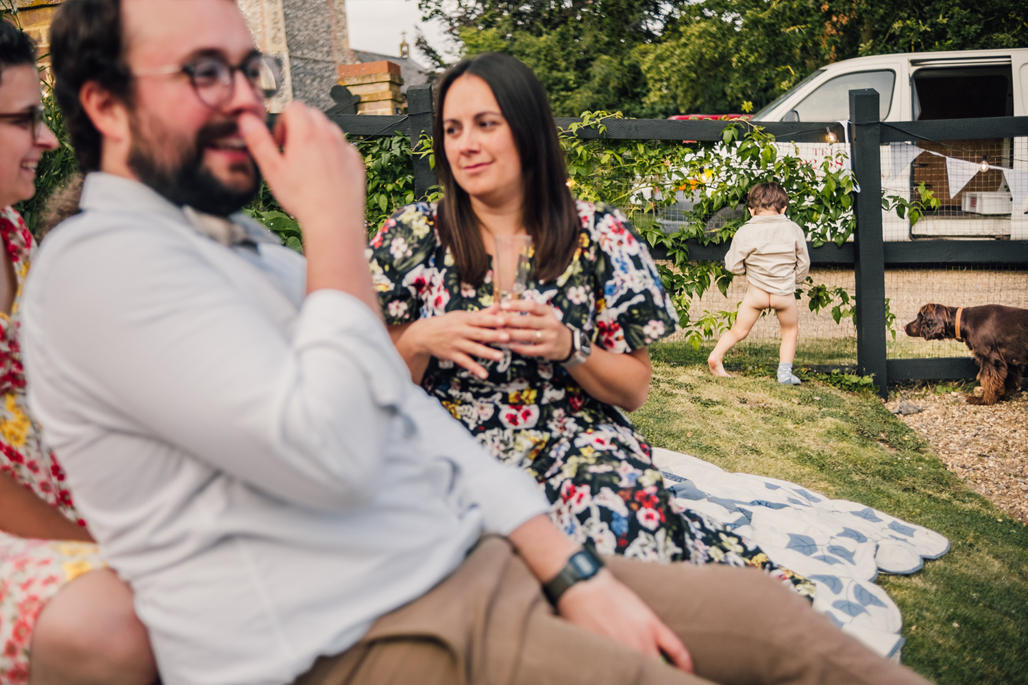 People sitting on a blanket in a backyard, socializing, with a child and dog playing near a fence in the background.