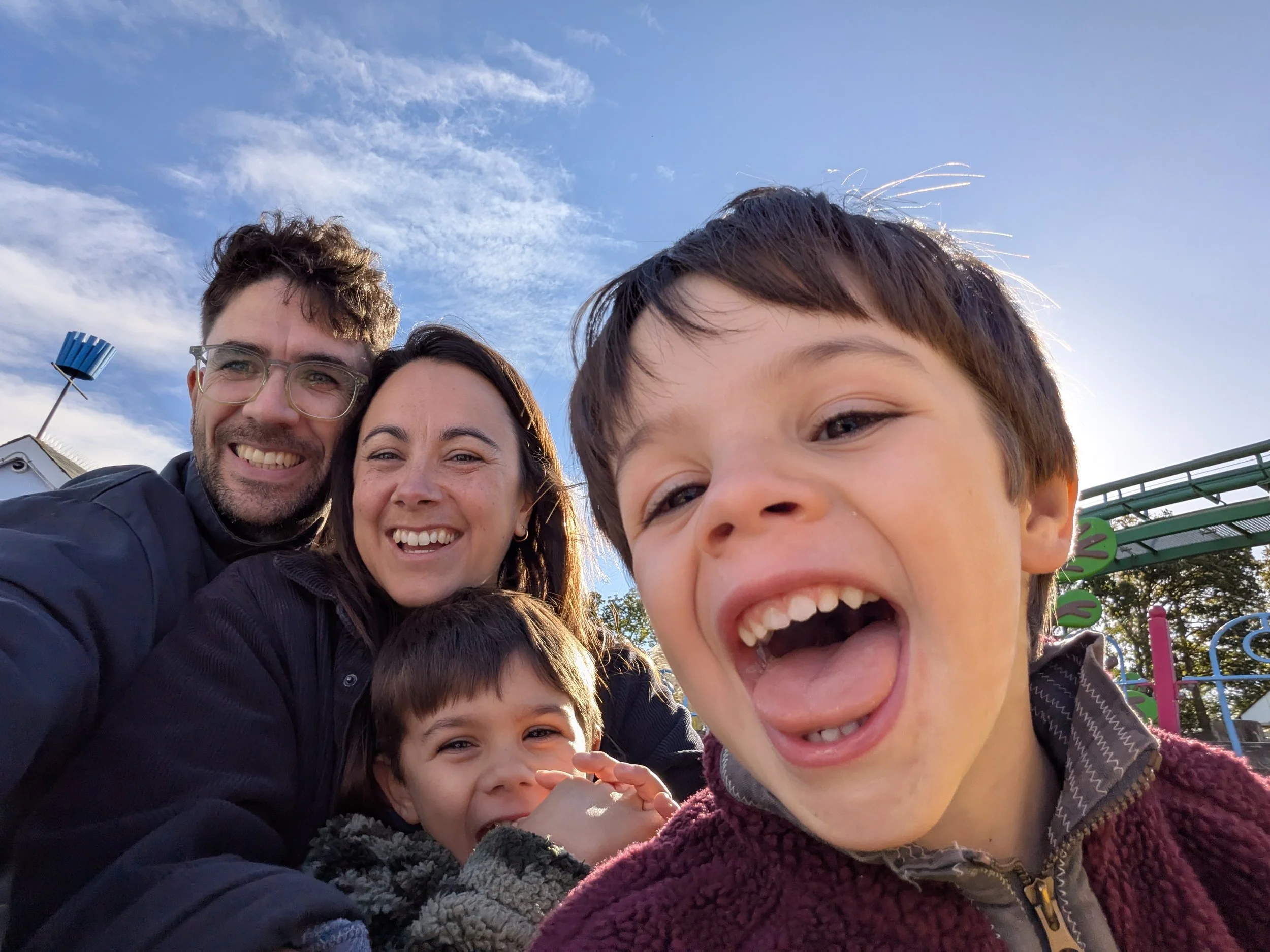 A group of five people, two adults and three children, taking a selfie outdoors during the daytime with a bright blue sky and some clouds in the background. They are smiling and appear to be enjoying themselves at a park or playground.