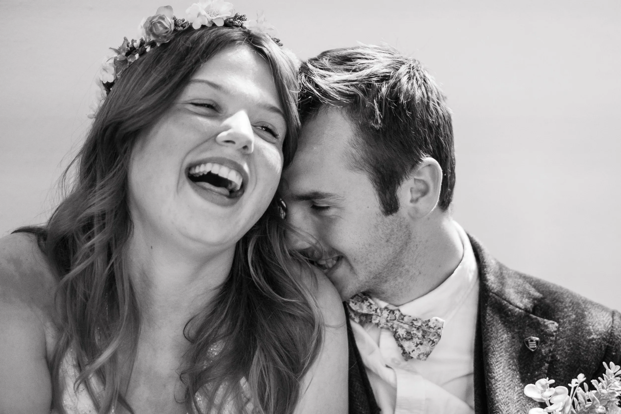 A black and white photo of a smiling woman with a flower crown and a man with short hair leaning in close, both appearing happy and affectionate.