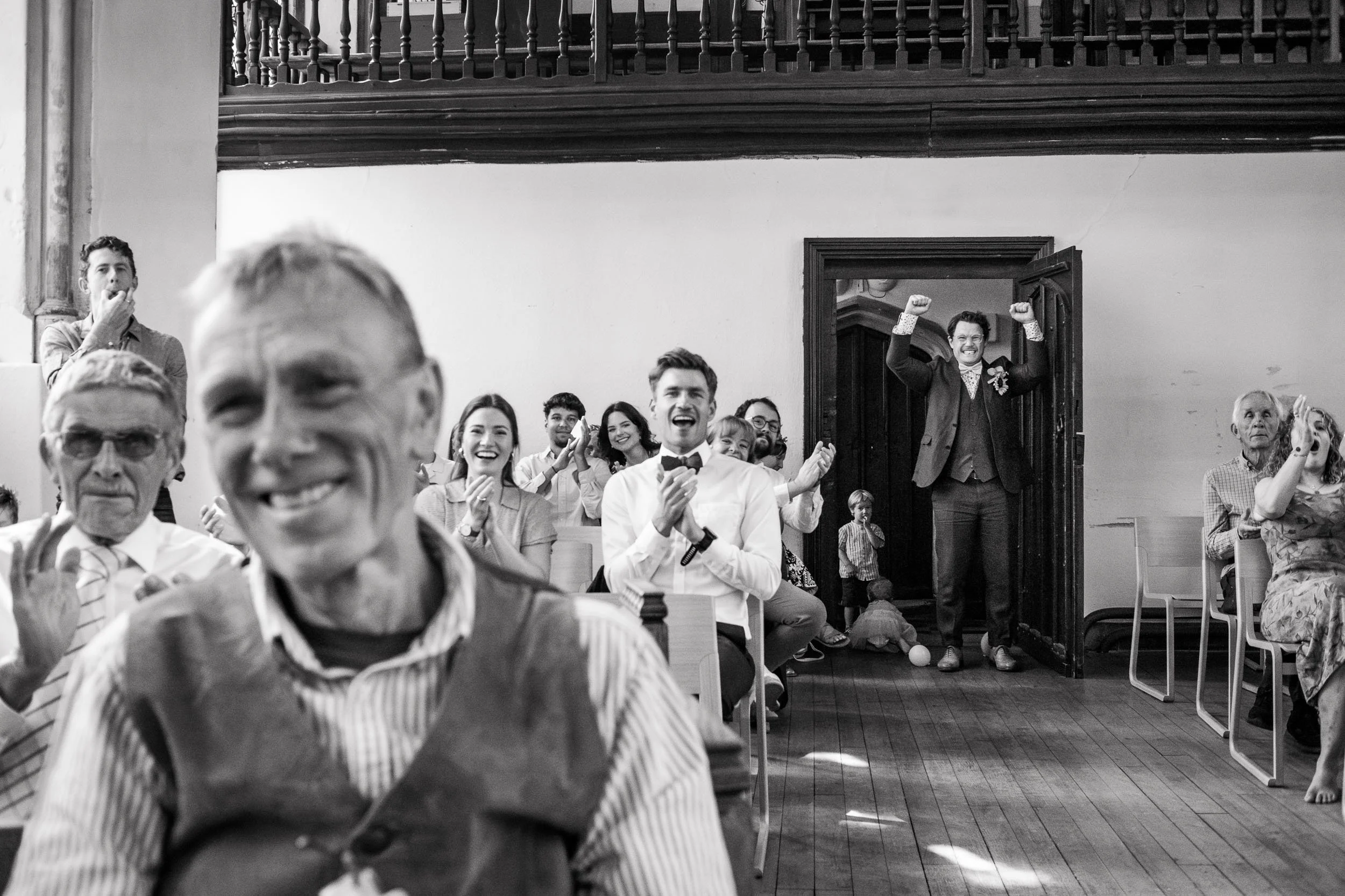 Group of people at a celebration, with a man in a tuxedo standing in a doorway with raised fists, others clapping and smiling, in a room with wooden floors and walls