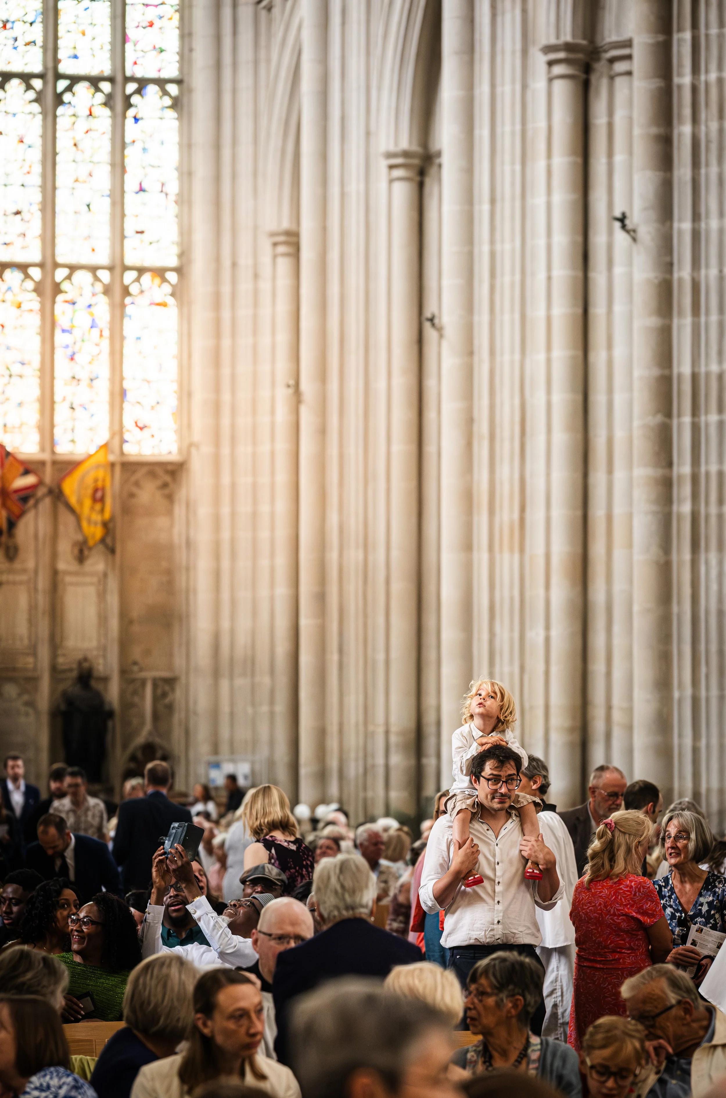 A man is carrying a young girl on his shoulders inside a grand church or cathedral with tall stained glass windows and stone columns, surrounded by a crowd of people.