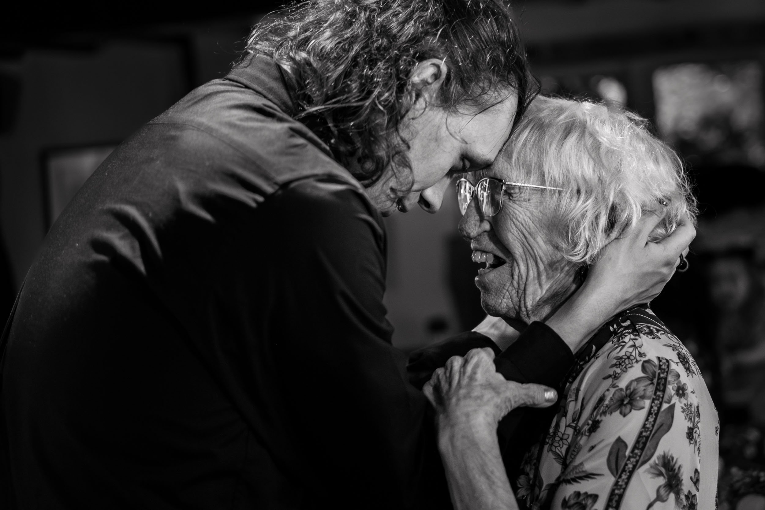 A young man and an elderly woman share an emotional hug, touching foreheads and smiling.