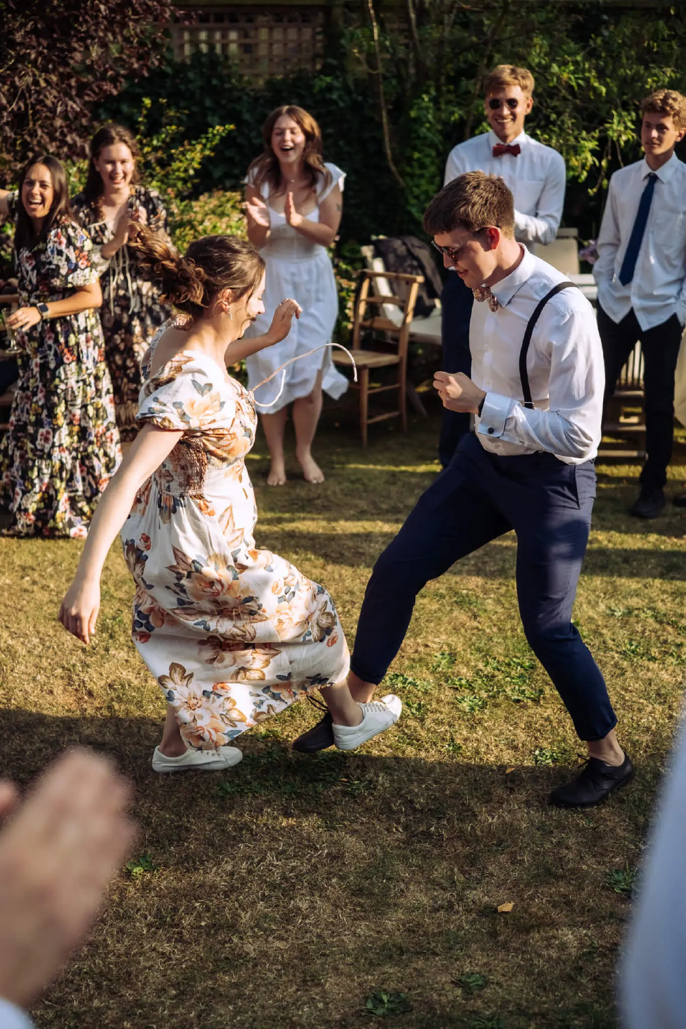 A group of friends dancing and laughing outdoors in a garden, with some people clapping and watching.