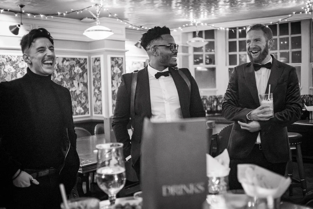 Three men in formal attire, two wearing tuxedos and one wearing a turtleneck, happily conversing and laughing at a dinner event in a decorated restaurant or banquet hall.