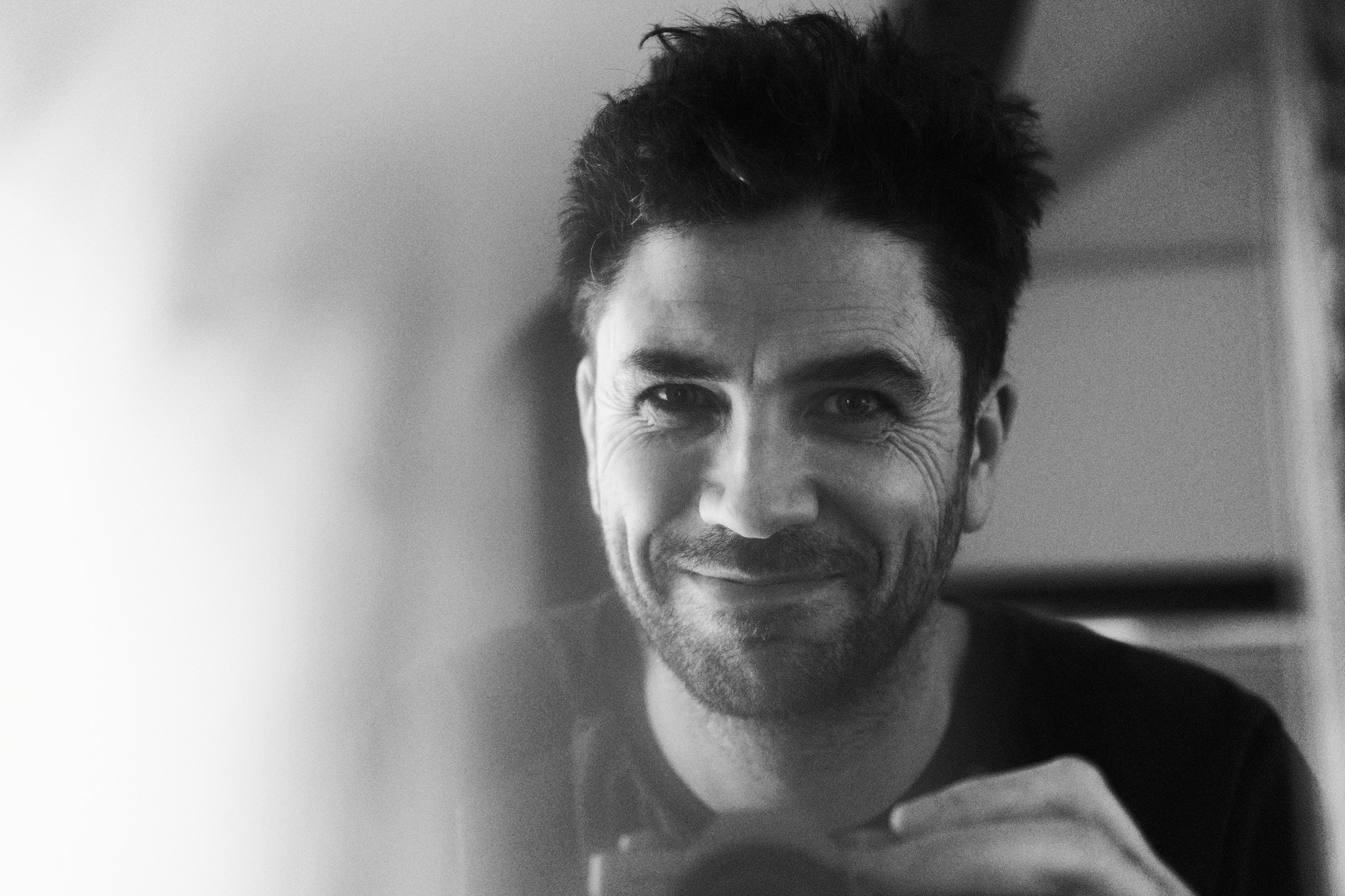 Black and white close-up photograph of a smiling man with dark, tousled hair, wearing a dark shirt, in front of a mirror holding a camera to take a selfie.