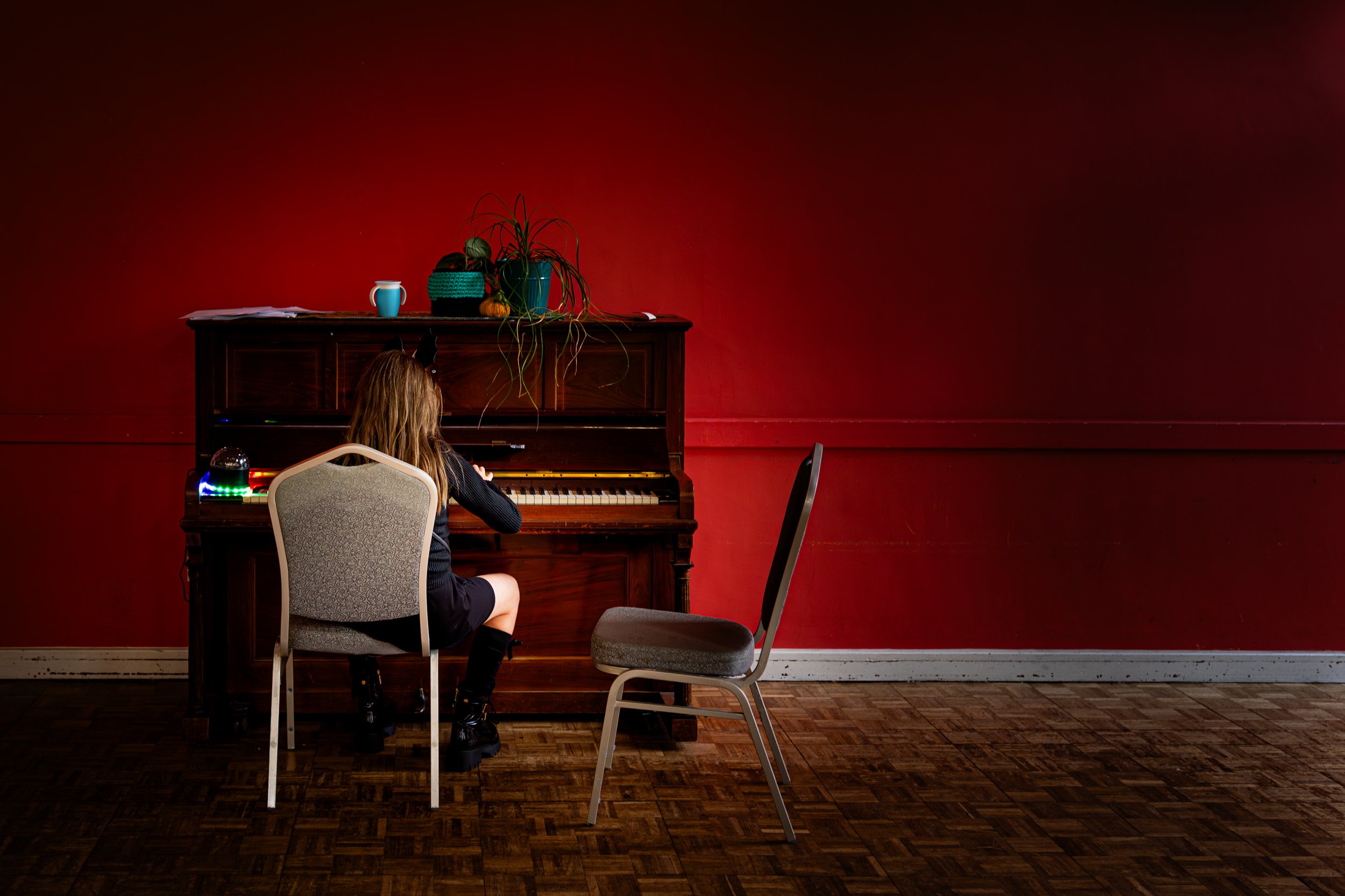 A girl with long hair sitting in a chair and playing an upright piano against a red wall.