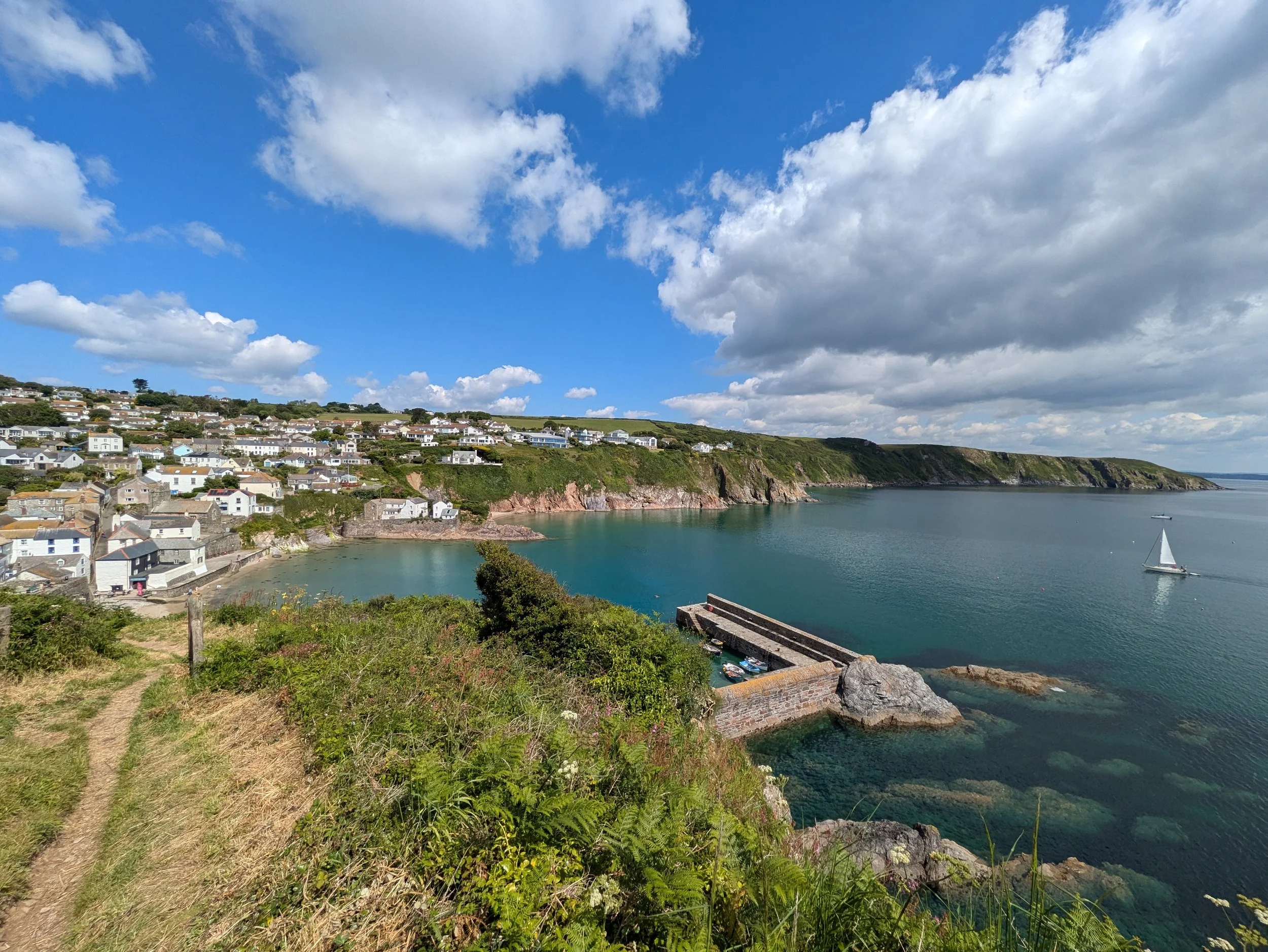 A coastal village with white houses on a hillside, overlooking a calm bay with a sailboat, under a partly cloudy sky.