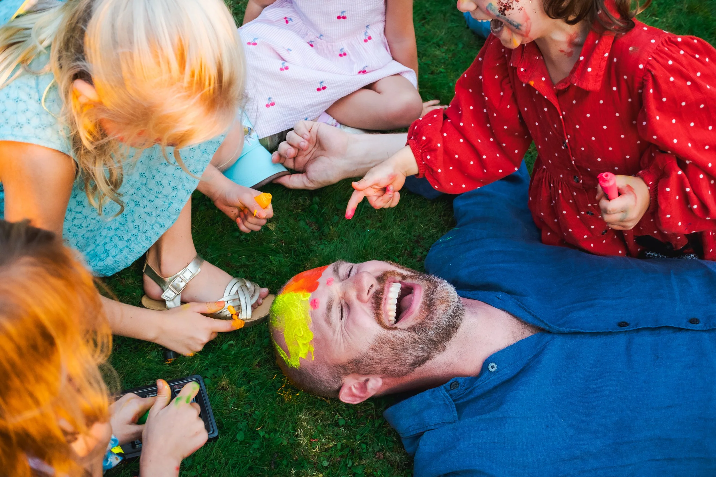 A group of children and a man with painted face, lying and sitting on grass, playing with chalk and laughing.