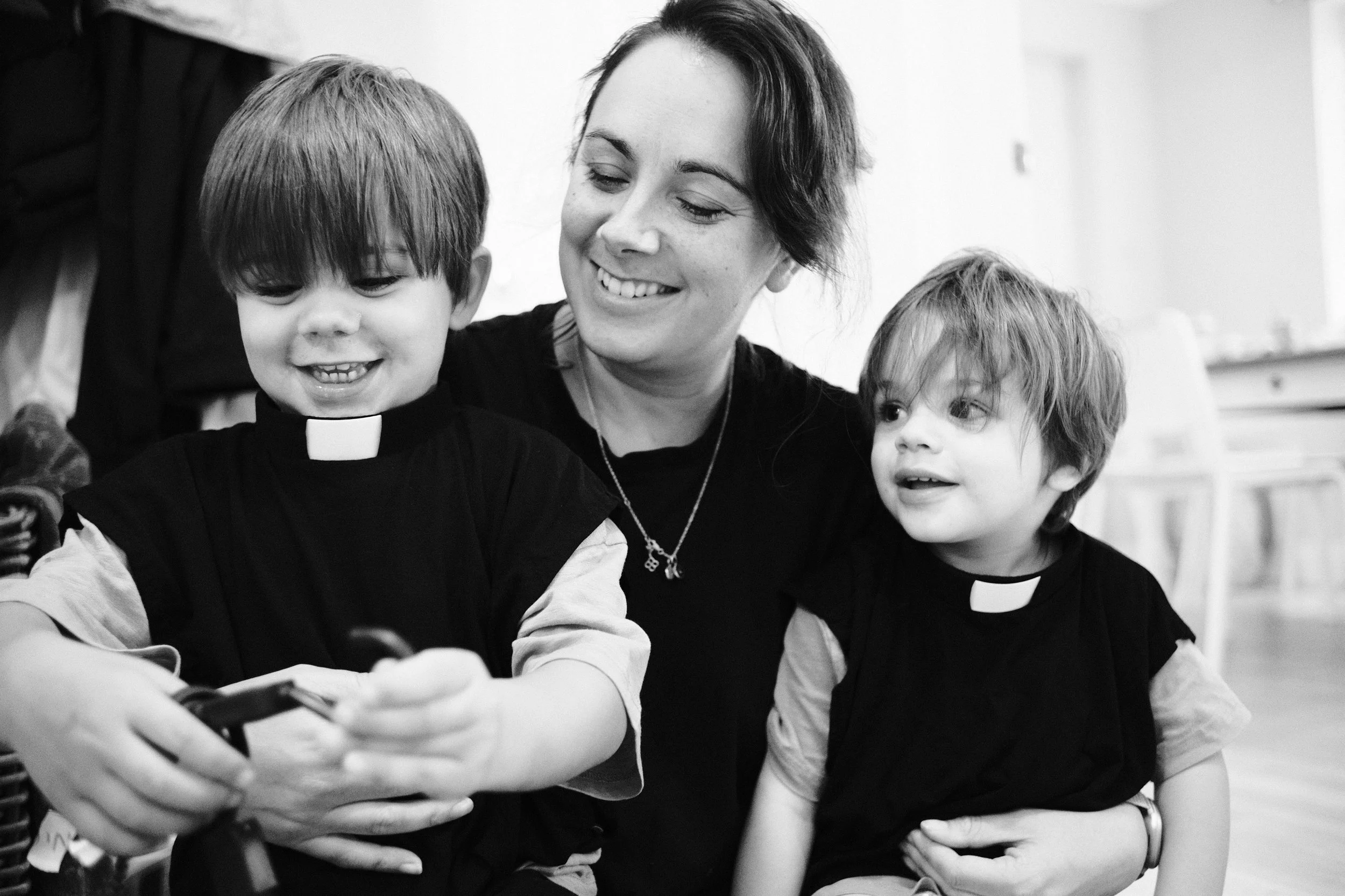 A woman dressed as a priest interacts with two young boys wearing religious attire, smiling and looking at a mobile device.
