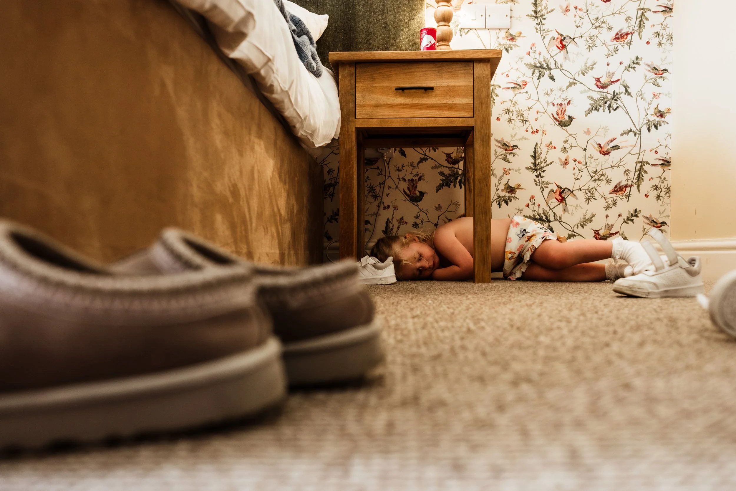 A young girl lies on the carpeted floor under a wooden bedside table, resting her head on the floor and smiling. The scene is viewed from a low angle, with sneakers and slippers in the foreground and a floral wallpaper background.