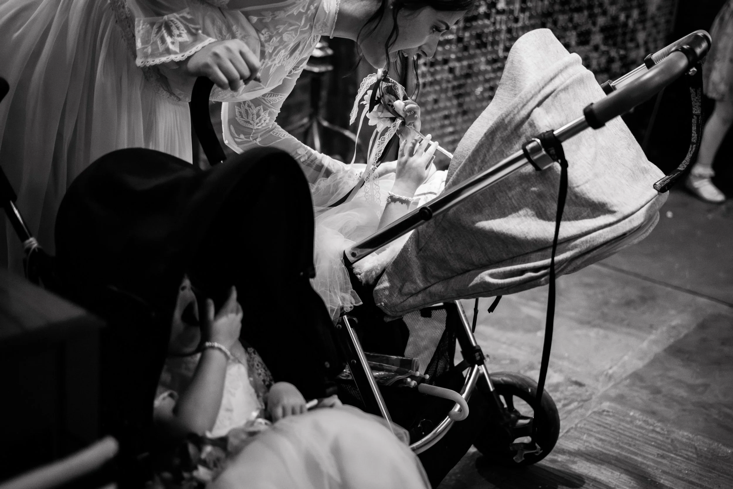 A young girl in a lace dress is administering an injection to a person lying in a medical stroller. Another person, possibly a child, is sitting nearby. The scene is in black and white.