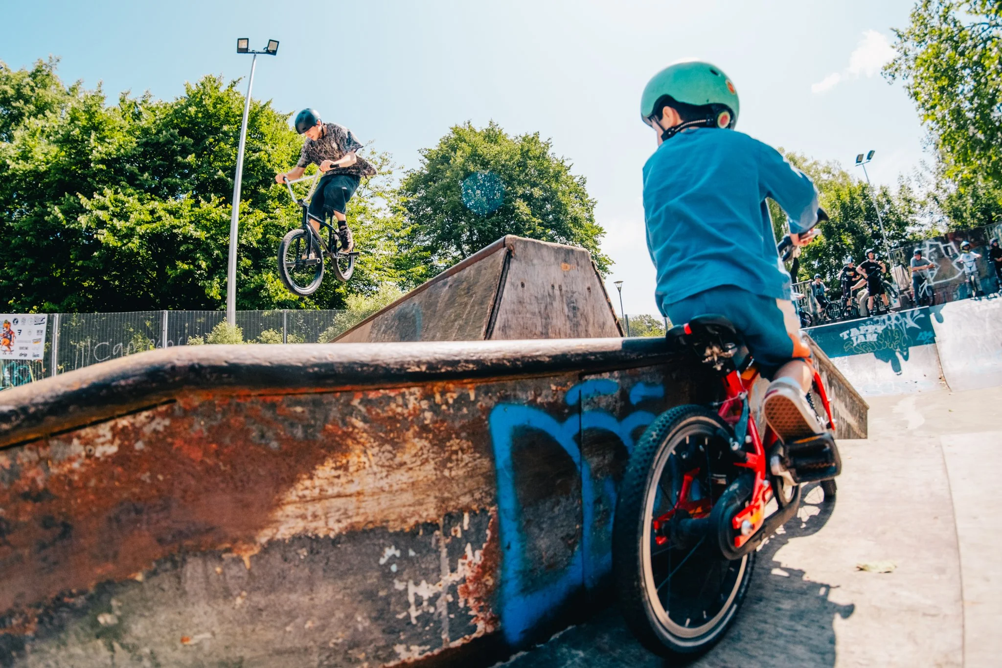 Children riding bikes at a skate park during daytime, with one child jumping off a ramp and another riding on the ground, surrounded by trees and spectators.