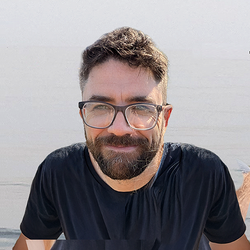 A man with glasses and a beard smiling outdoors near water.