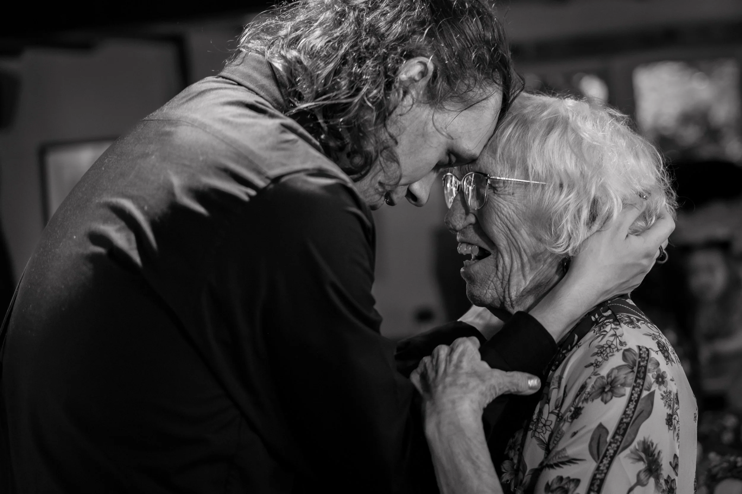 A young man and an elderly woman share an emotional hug, touching foreheads and smiling.