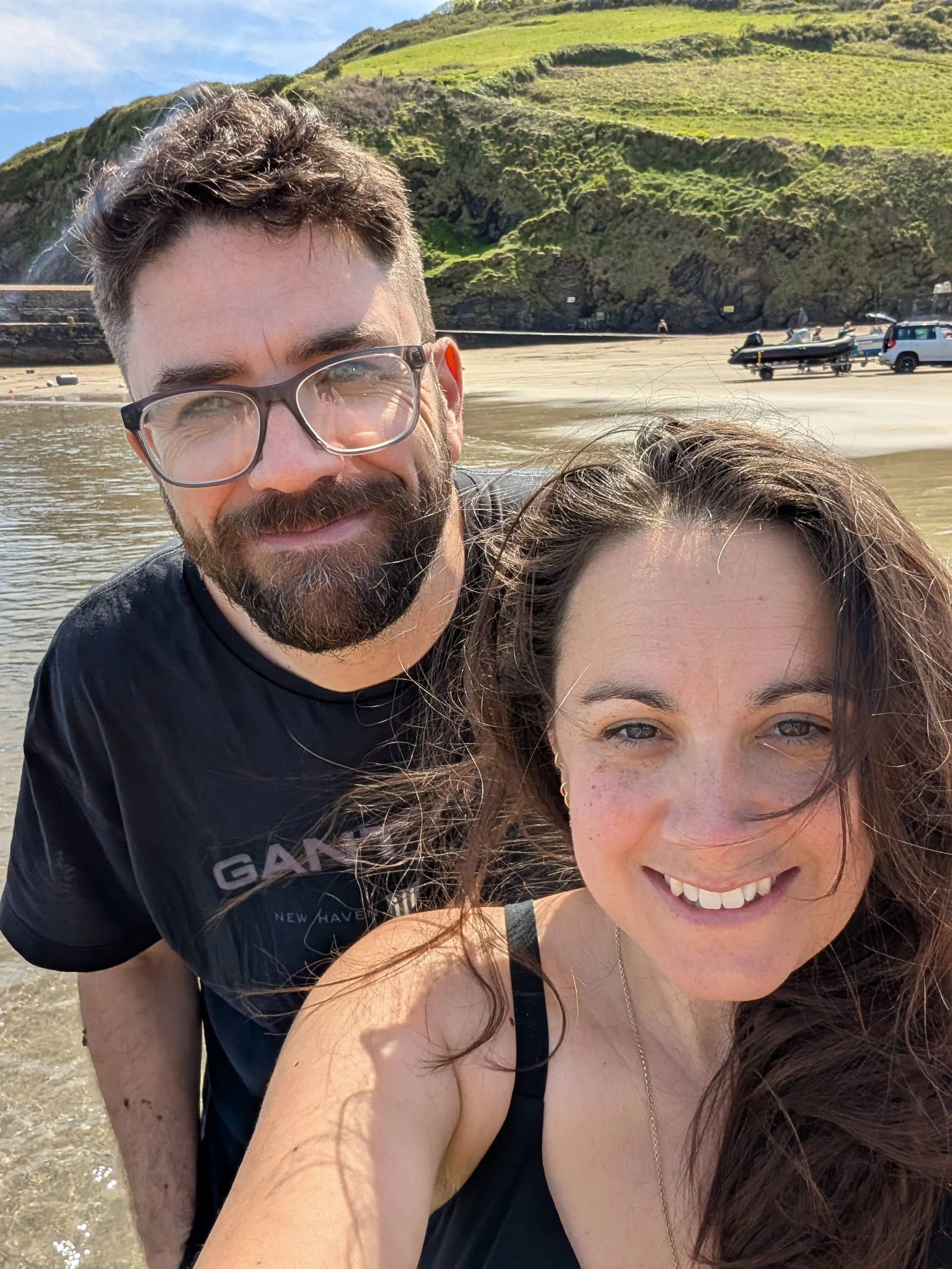 Smiling woman with curly brown hair and a man with glasses and a beard, both at the beach with green hills and a boat in the background.