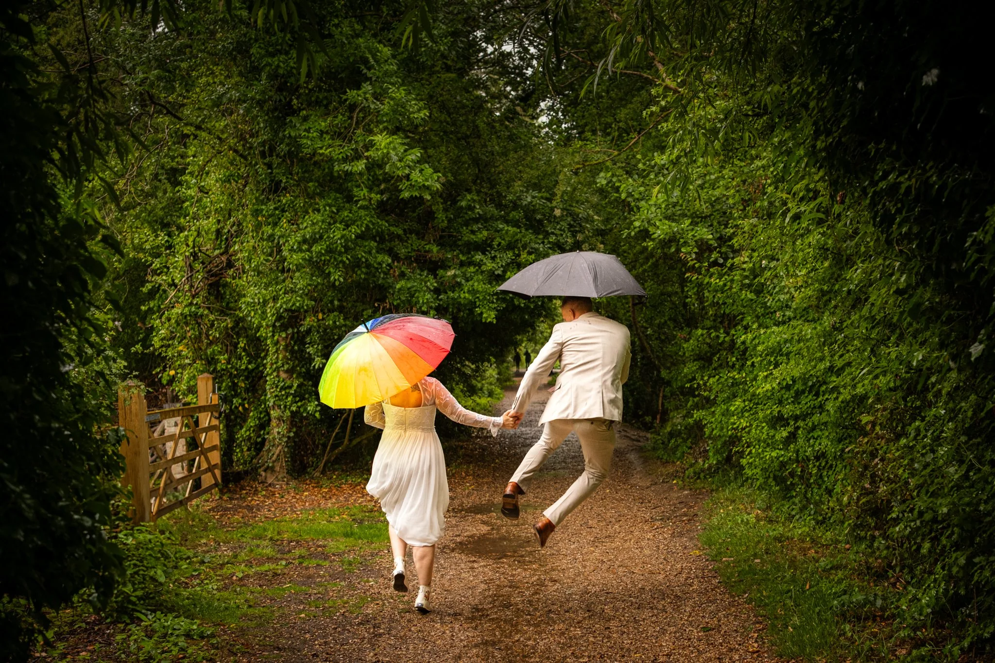 A couple dressed in wedding attire holding hands and jumping on a rainy day with umbrellas in a lush green forested path.