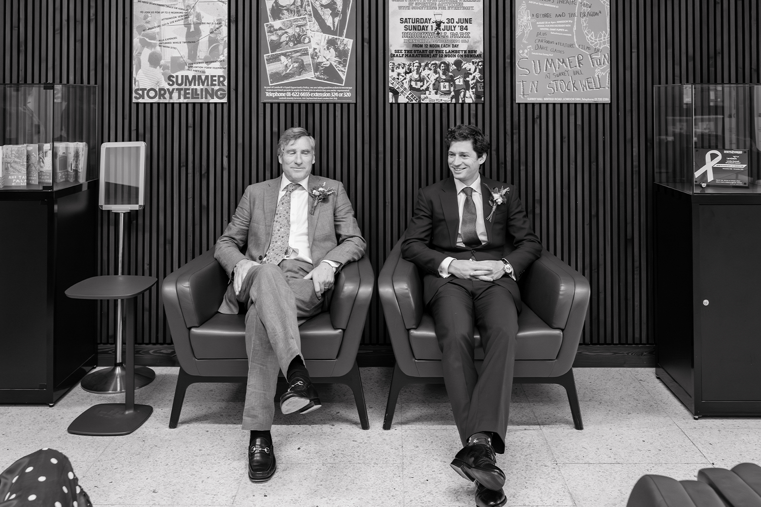 Two men in suits sitting in armchairs inside a building with wood-panel walls, smiling. Posters are on the wall behind them, and a display case is on each side.