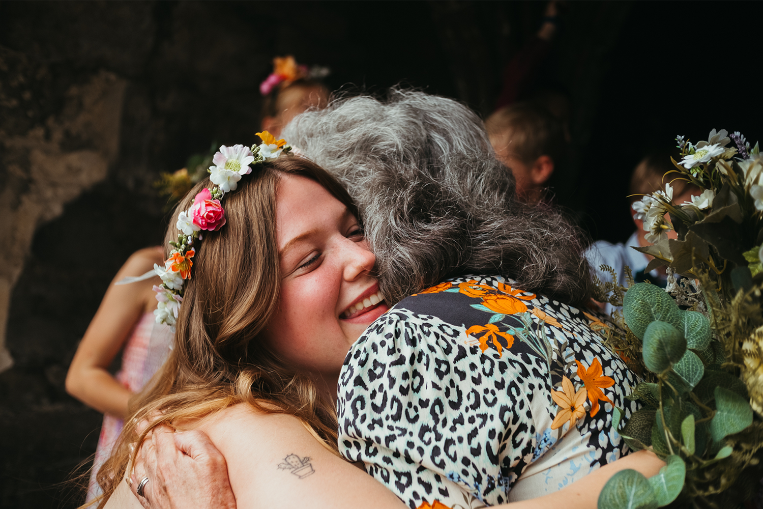 Young woman with a flower crown hugging an older woman, smiling with eyes closed, surrounded by others at a celebration, possibly a wedding or festival.