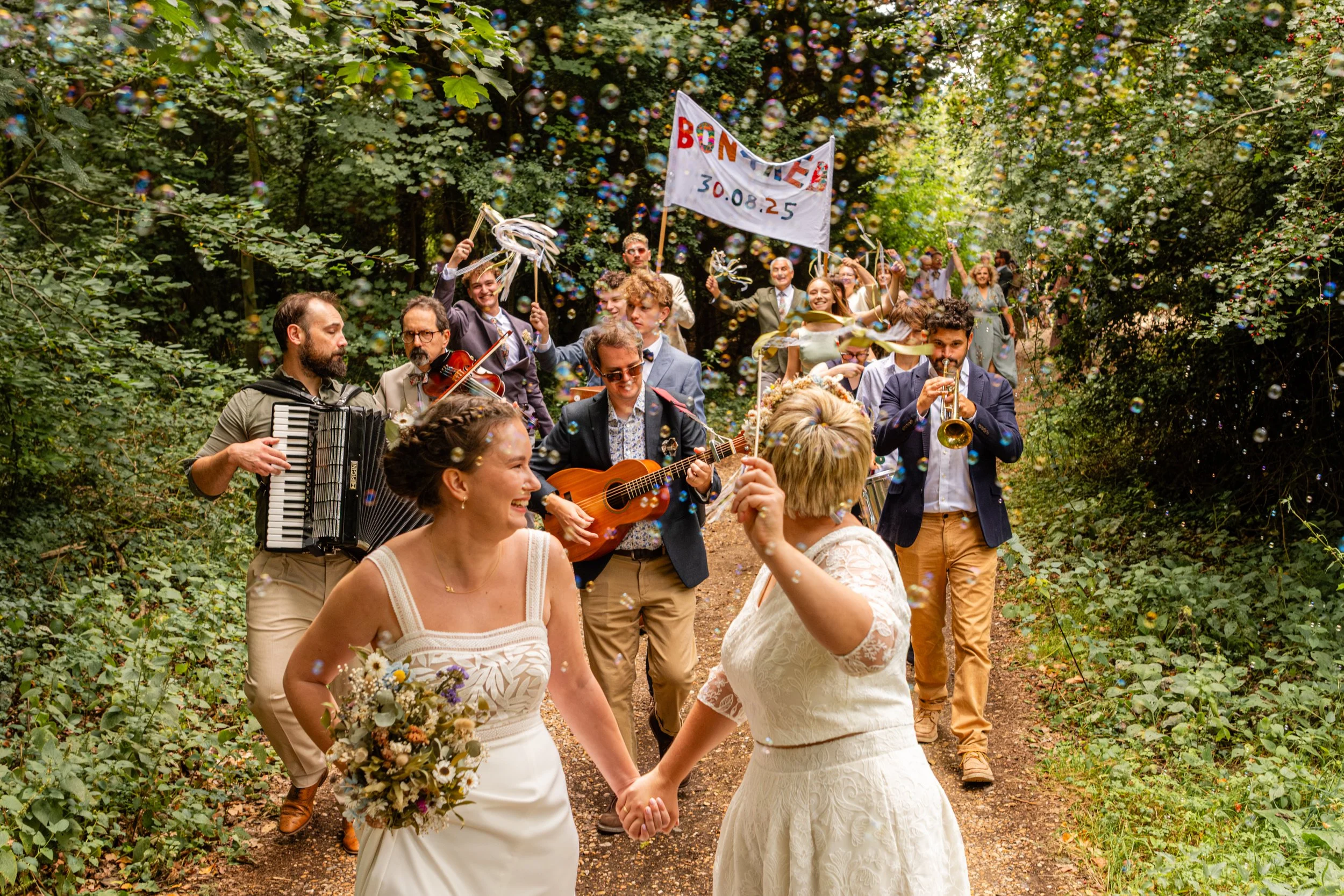 A wedding celebration with two brides holding hands and dancing in front of a group of musicians playing guitar, accordion, violin, and trumpet in a wooded outdoor setting, with bubbles, a colorful banner, and joyful guests.