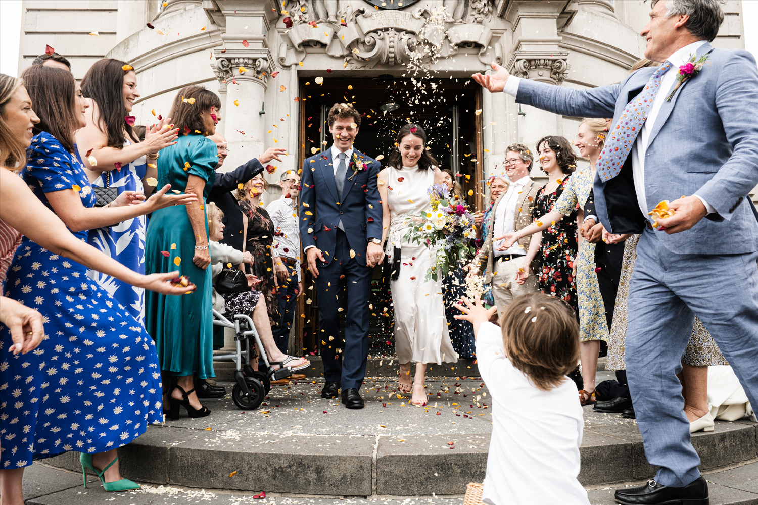 A newly married couple walking out of a church surrounded by family and friends throwing flower petals and confetti celebrating their wedding.