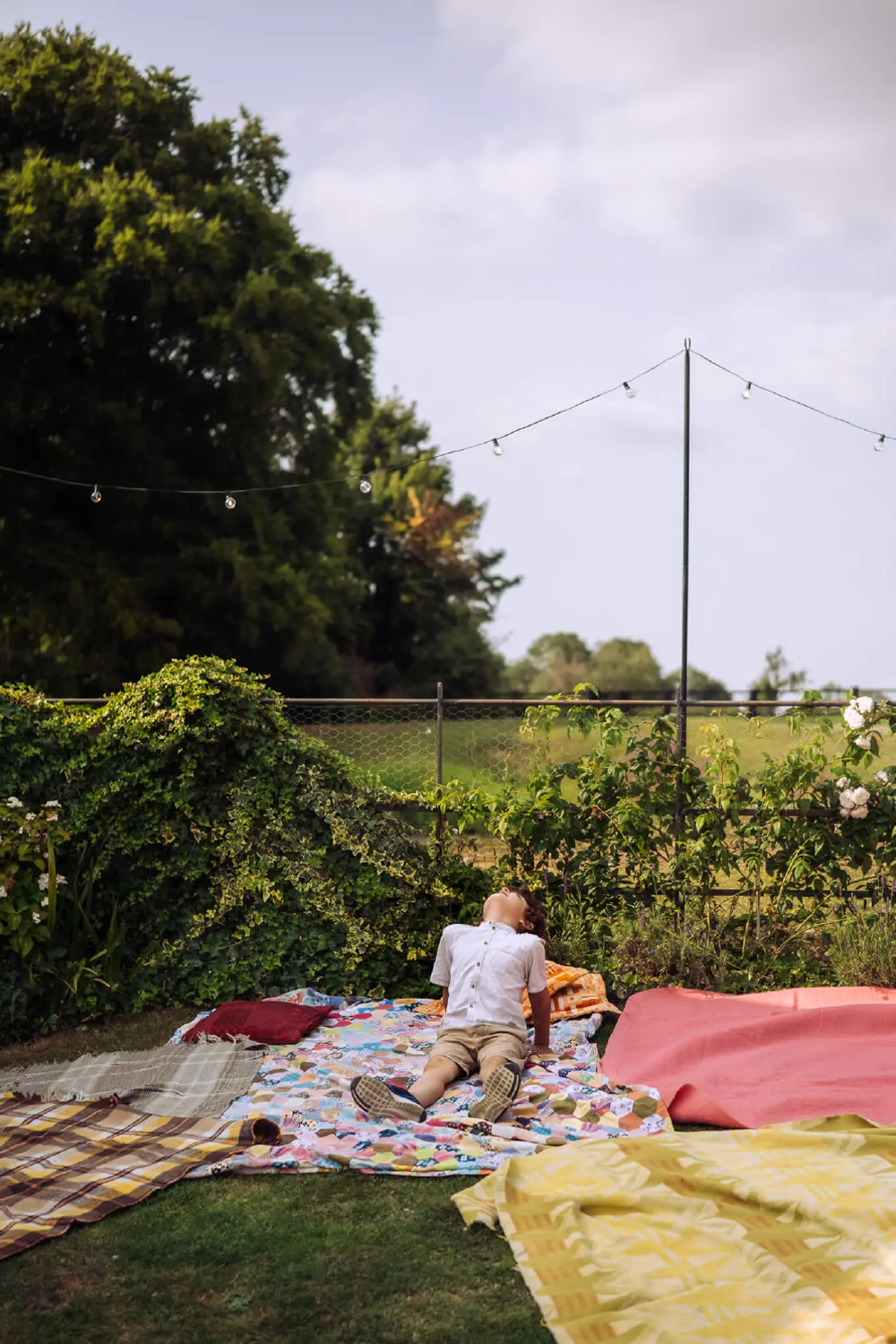 Child lying on a colorful quilt outdoors in a garden, surrounded by blankets and pillows, looking up at the sky with string lights overhead, greenery and trees in the background.