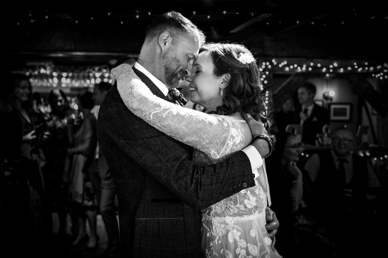 A couple dancing closely at their wedding reception, with the groom in a suit and the bride in a lace dress, surrounded by friends and family in a decorated indoor venue.