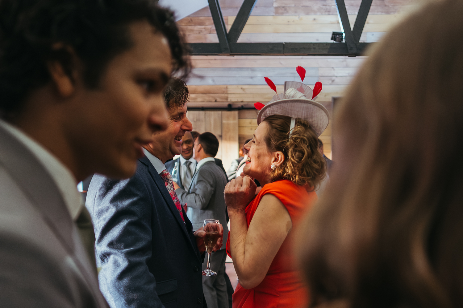 A group of people at a social gathering, with a woman in a bright orange dress and large hat decorated with red elements, smiling and laughing with a man in a suit, while other guests converse in the background, in a wooden interior space.