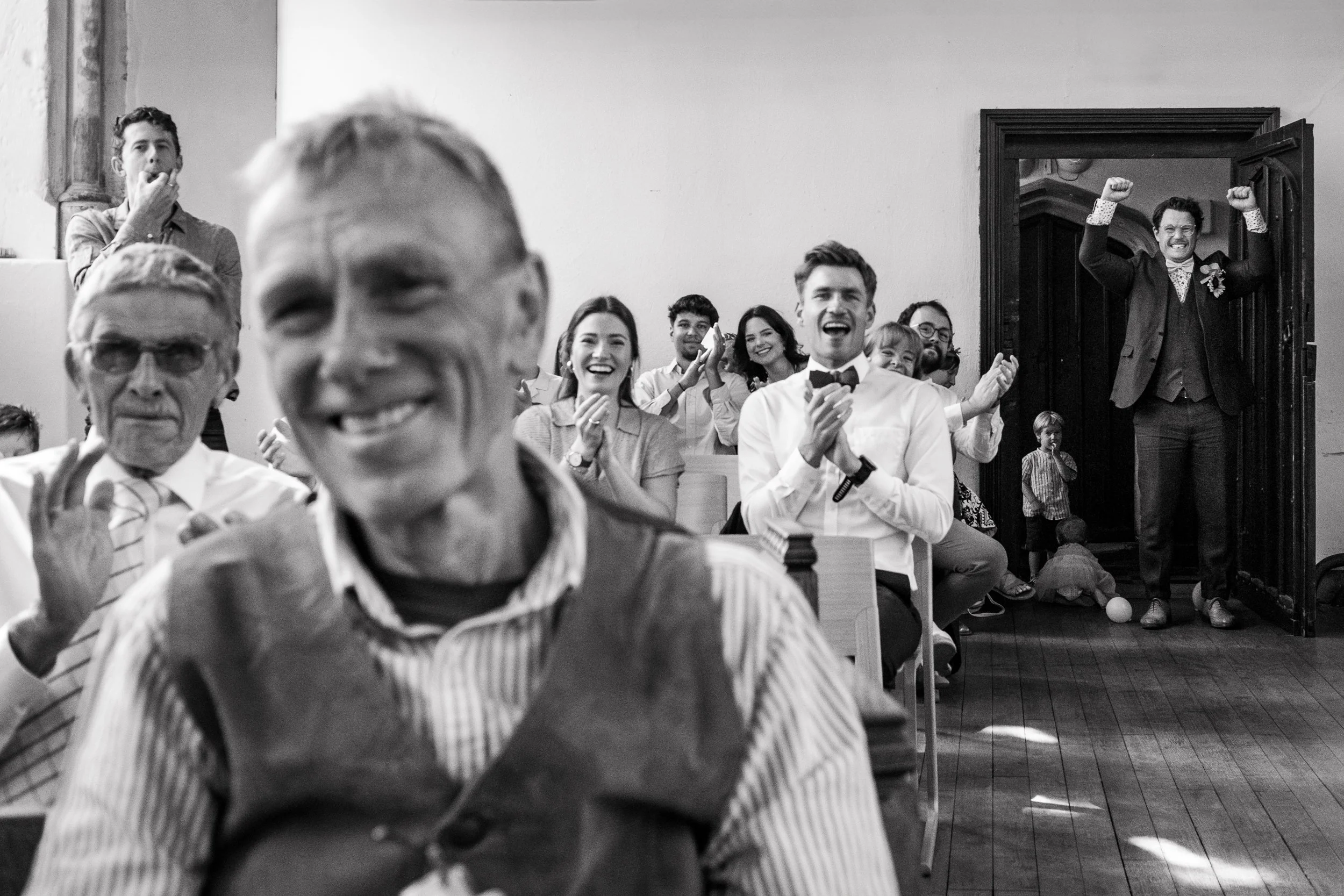 Group of people at a celebration, with a man in a tuxedo standing in a doorway with raised fists, others clapping and smiling, in a room with wooden floors and walls