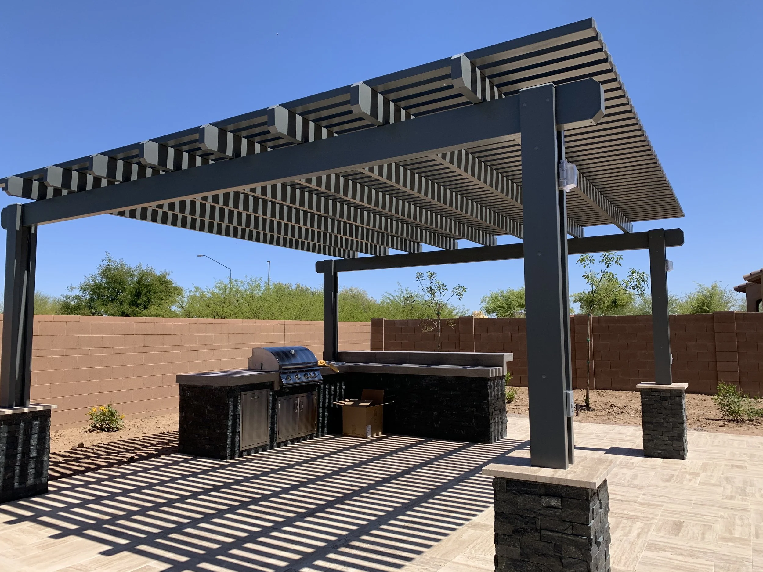 Outdoor patio area with a grill, black stone counter, and a pergola with striped shade, in a backyard with a brick wall and small trees.