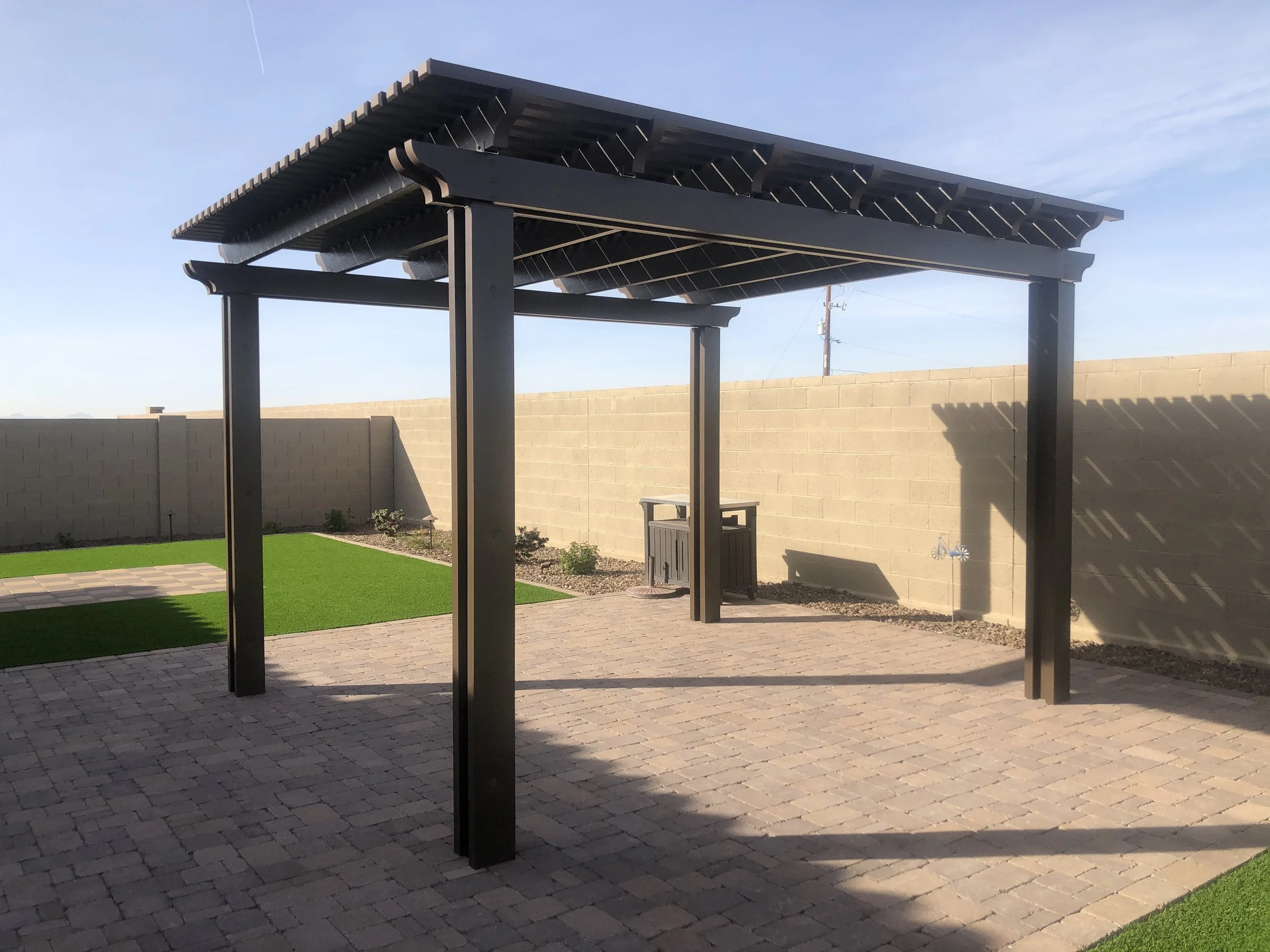 Backyard patio featuring a black pergola over brick pavers, with a small green grass area, a tan brick wall, and a small decorative windmill. There's also a trash bin and a garden bed with plants.