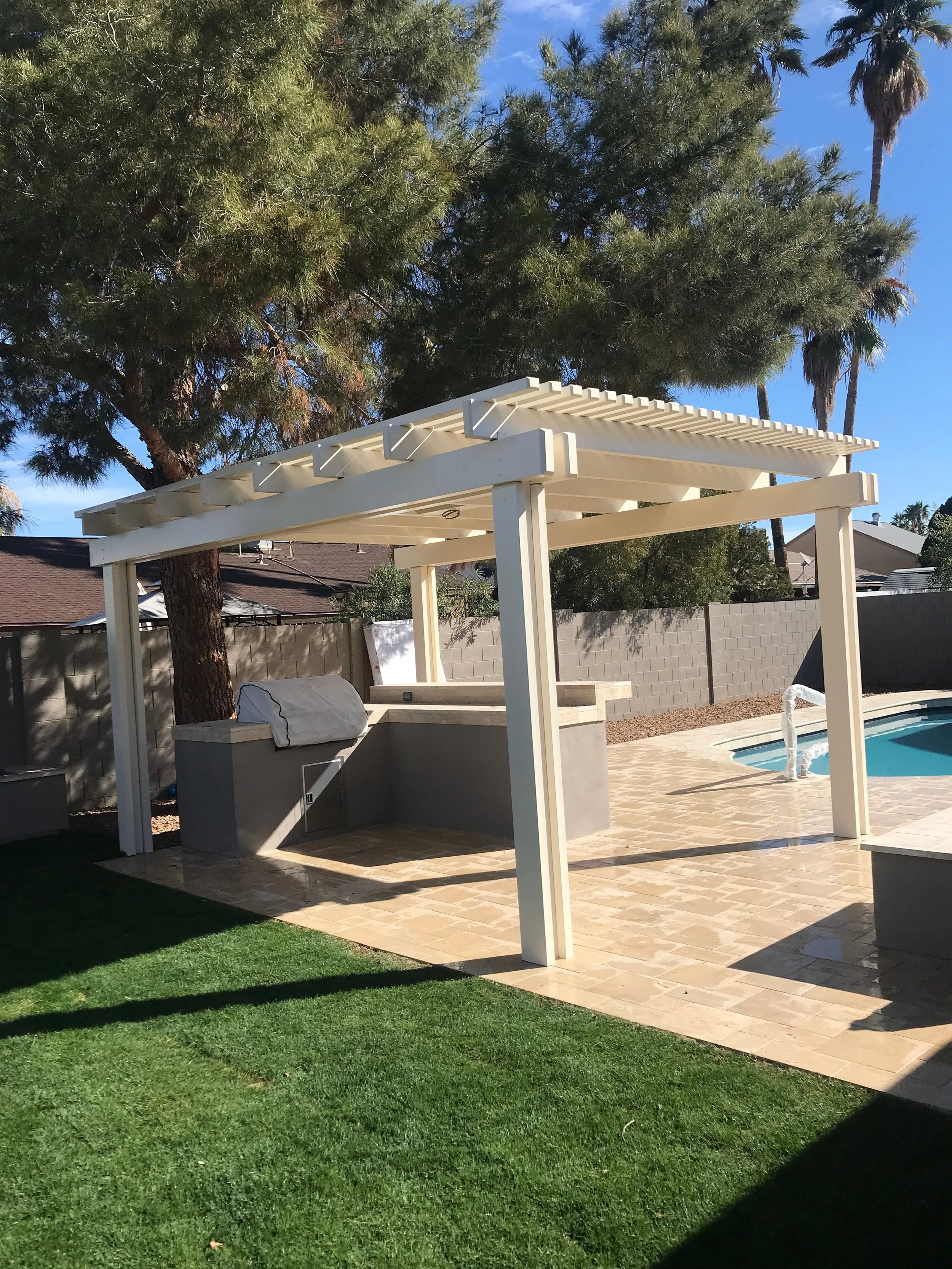 Backyard with a pool, covered patio area with a white pergola, gray built-in grill, stone patio, green grass, large trees, and a concrete block wall.