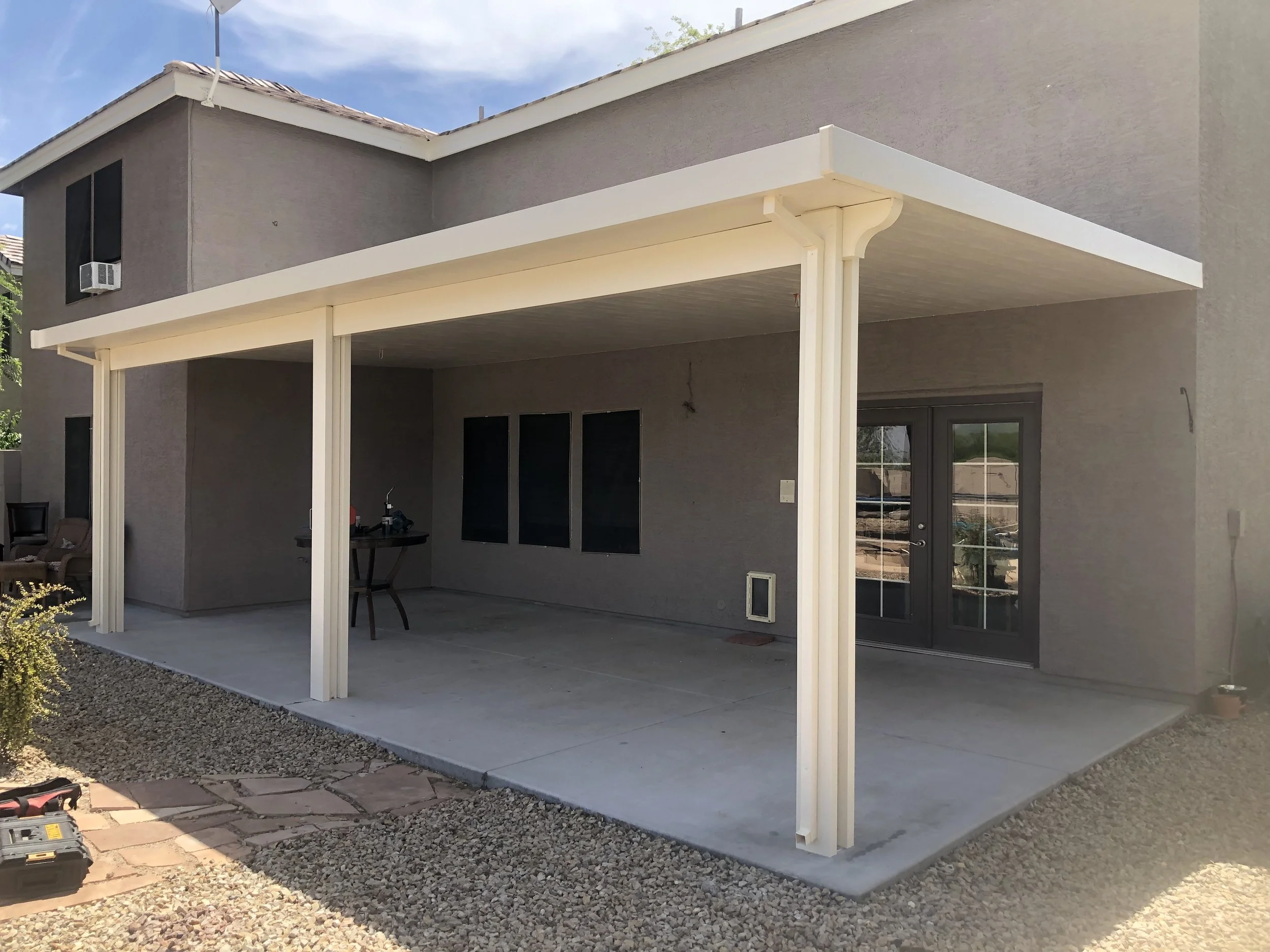 Backyard patio with a concrete floor, metal framing for a future roof, and a sliding glass door leading into the house. There are a few chairs and a small table on the patio.